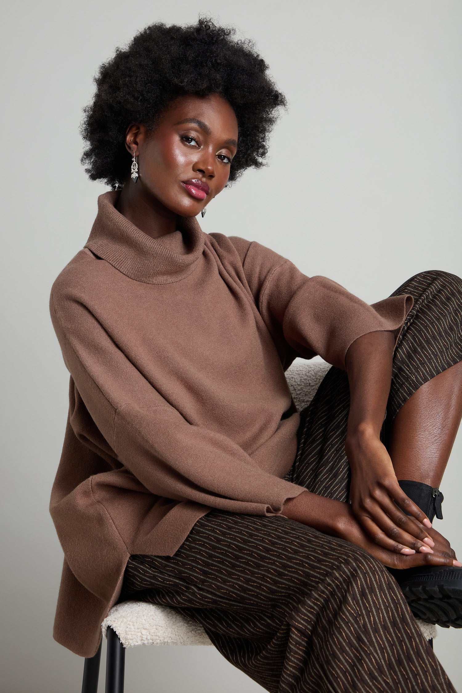 A seated individual is wearing a brown turtleneck sweater and eye-catching Mini Check Jersey Bubble Trousers. They complete the look with black boots, resting their elbow on a knee against a neutral background.