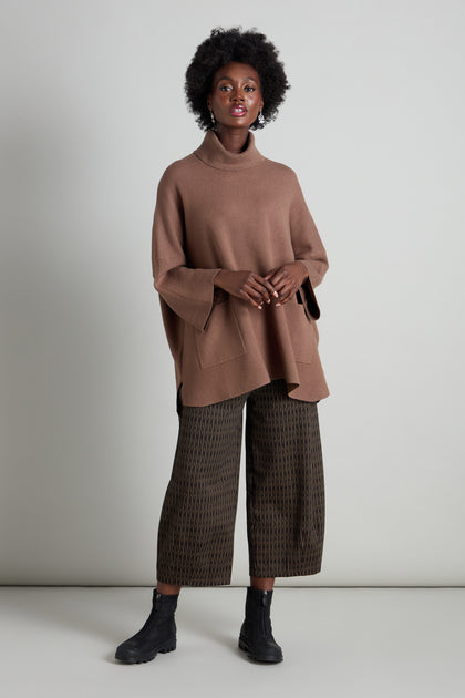 A person wearing a brown turtleneck poncho, mini check jersey bubble trousers, and black ankle boots stands against a plain background.