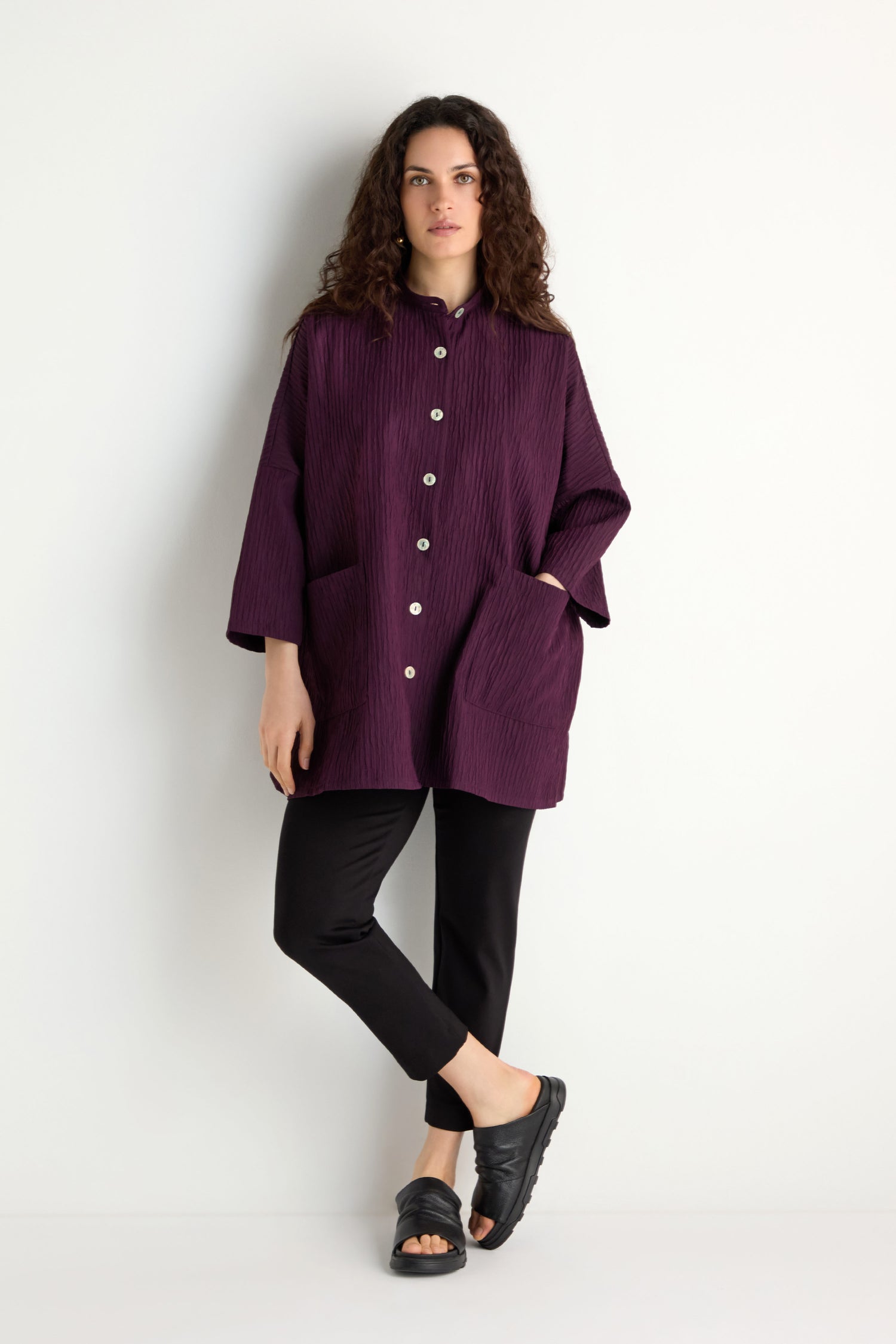A woman poses against a white background in the Pleated Pocket Jacket, dark purple, styled with black pants and sandals. Her curly hair frames her face as she stands with a neutral expression.