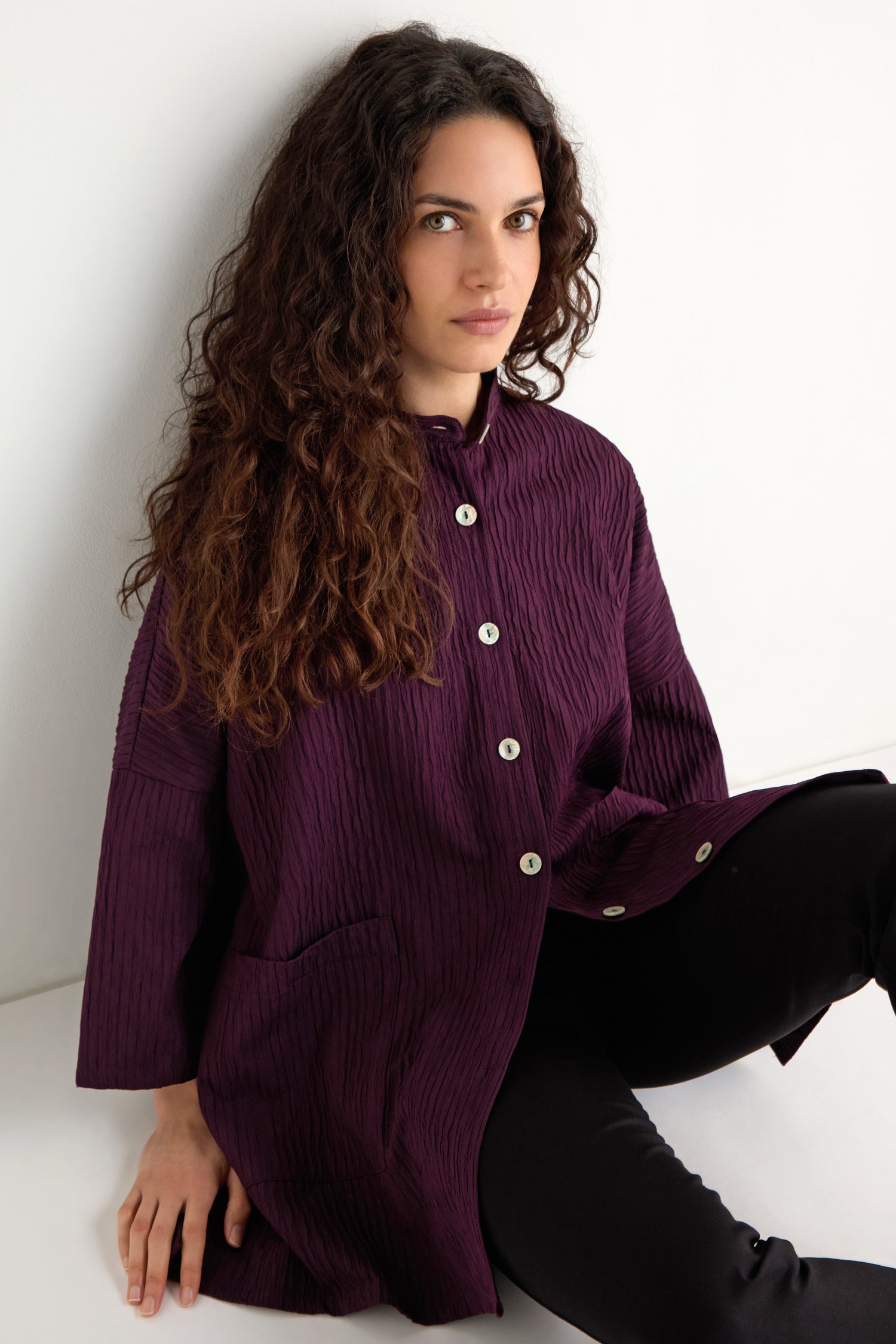 A woman with long curly hair wears the Pleated Pocket Jacket in purple with black pants, sitting against a white wall and looking at the camera.