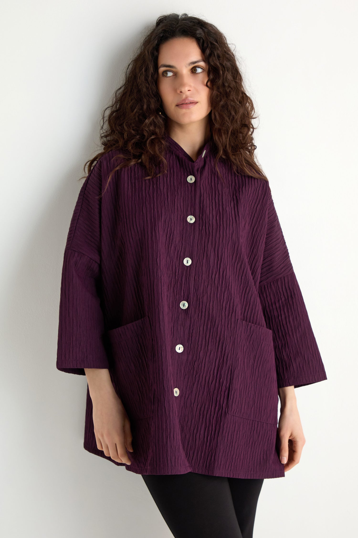 A woman with long curly hair wears the Pleated Pocket Jacket, a loose-fitting textured purple jacket with large front pockets, paired with black leggings, standing against a white background.