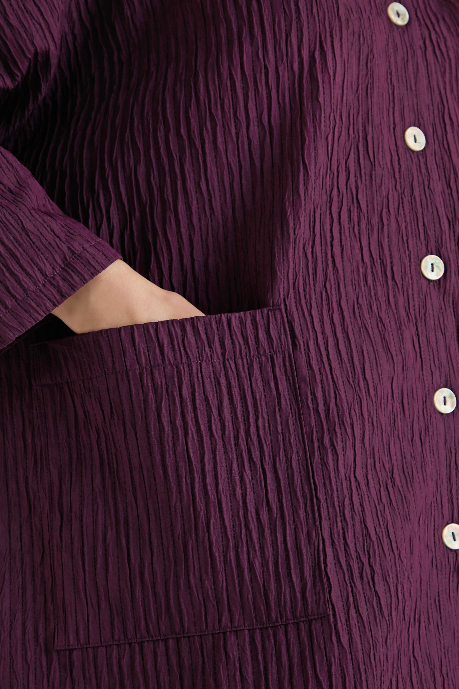 Close-up of a woman's hand in the pocket of the Pleated Pocket Jacket, featuring a plum color, white buttons, and crafted from three-dimensional pleated fabric.