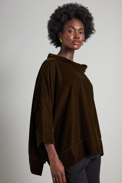 A person wearing a Cowl Neck Boxy Top in dark brown with 3/4 length flared sleeves and black pants, short curly hair, and small hoop earrings stands against a plain background.