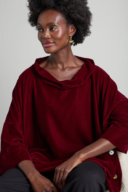 A person wearing a deep red Cowl Neck Boxy Top featuring 3/4 length flared sleeves and paired with black pants, sits gracefully, gazing to the side.