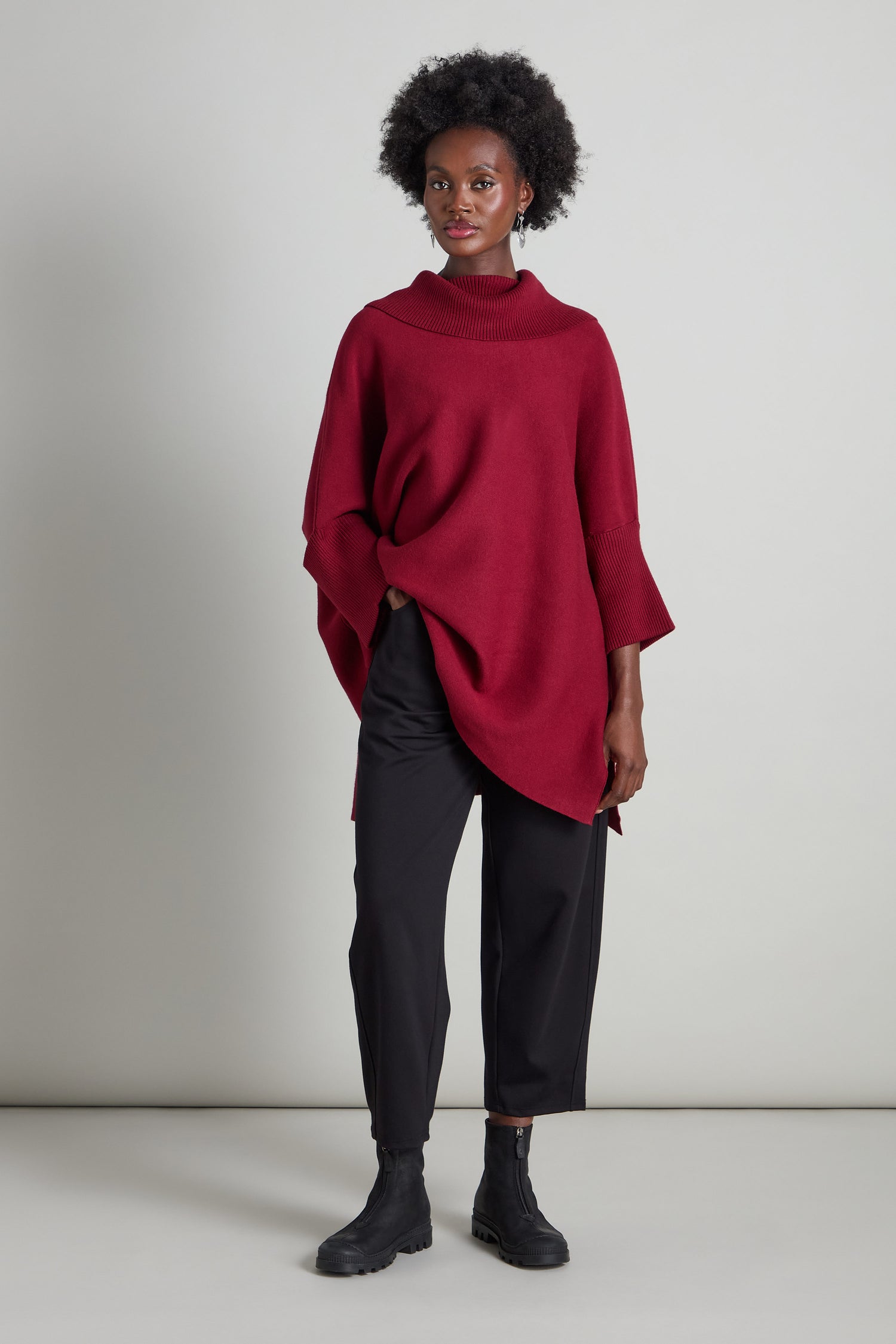 A person in a red turtleneck poncho stands against a gray background, effortlessly pairing it with black boots and the Slim Jersey Trouser that features a sleek slim-leg silhouette.