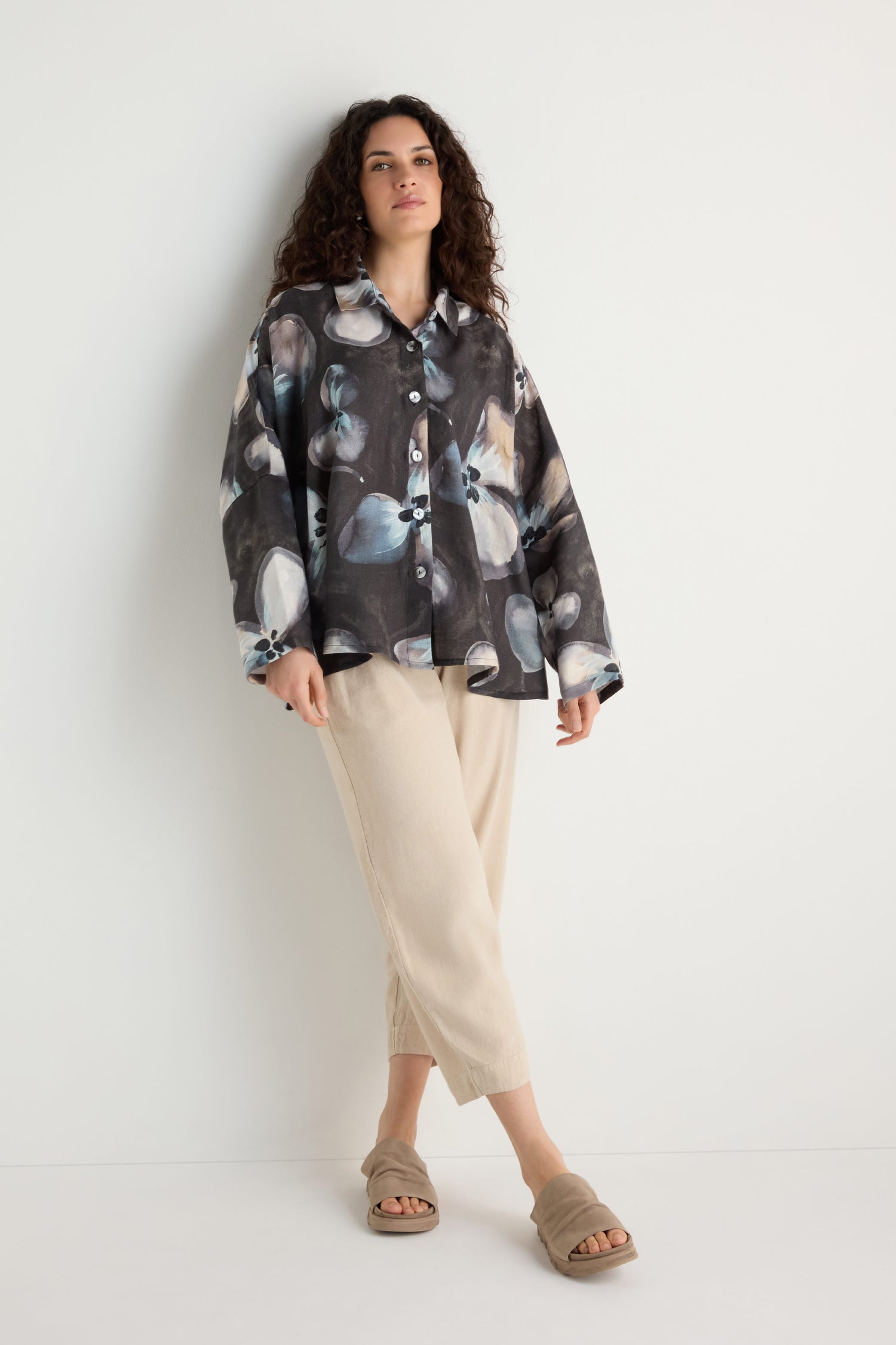 A person stands against a white wall wearing the Abstract Floral Linen Shirt, relaxed fit, paired with beige cropped pants and beige slide sandals.