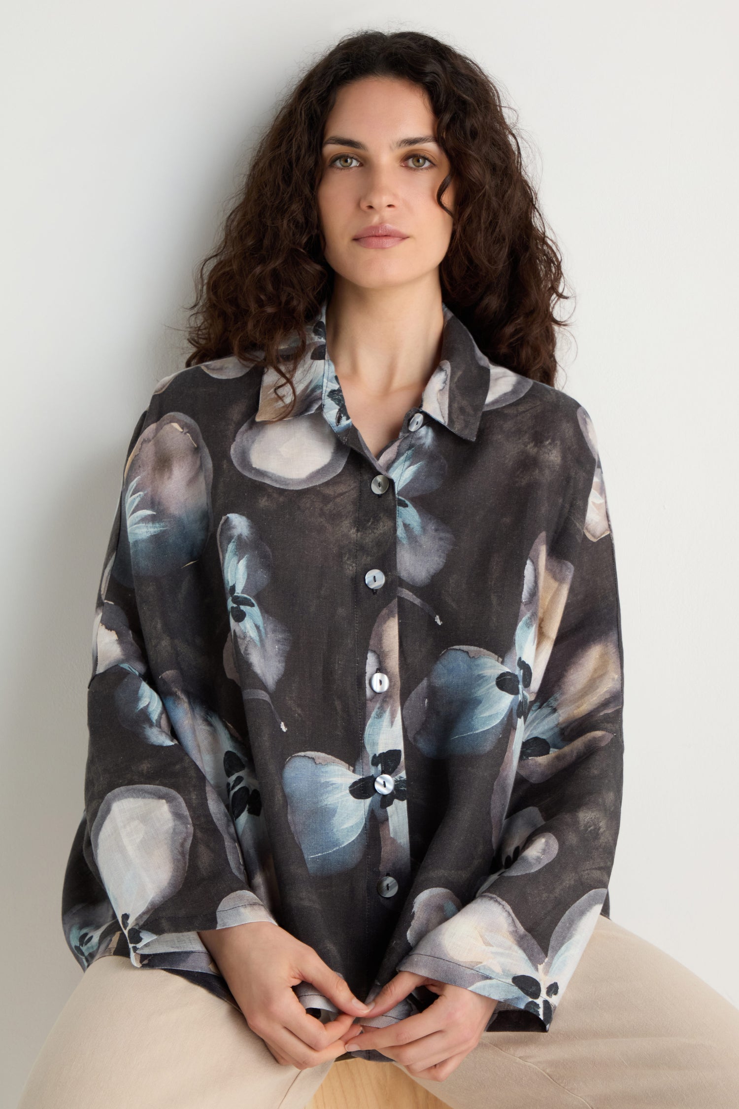 A woman with curly hair wears the Abstract Floral Linen Shirt, a relaxed fit with blue and gray abstract floral pattern, as she sits against a plain light background.
