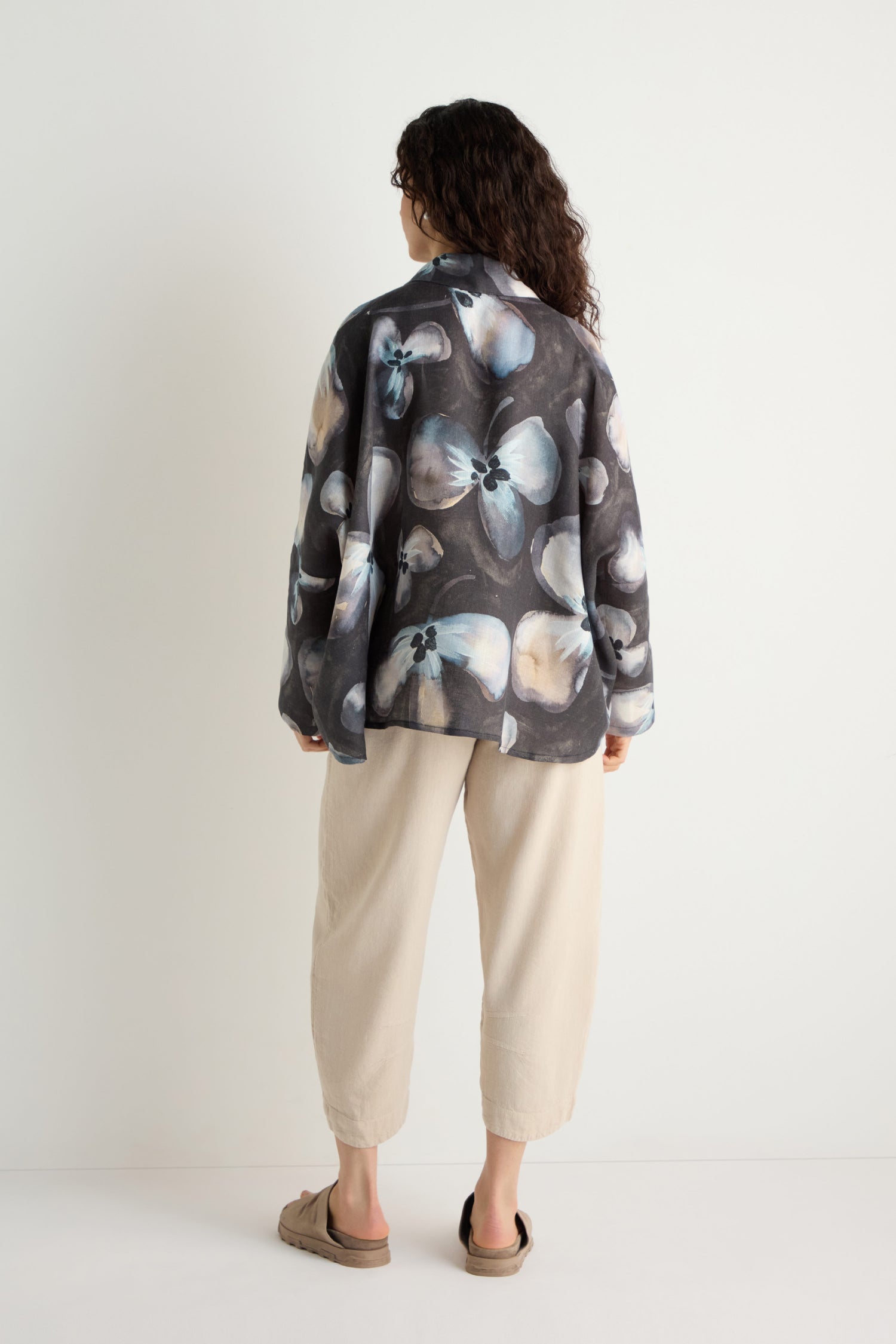 A person stands facing away, wearing the Abstract Floral Linen Shirt—relaxed fit with an abstract floral pattern—paired with light loose pants and sandals against a white background.