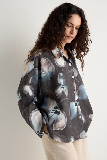 A woman with long curly hair wears the Abstract Floral Linen Shirt—relaxed fit in dark linen with an abstract floral pattern—paired with light pants, standing against a plain background.