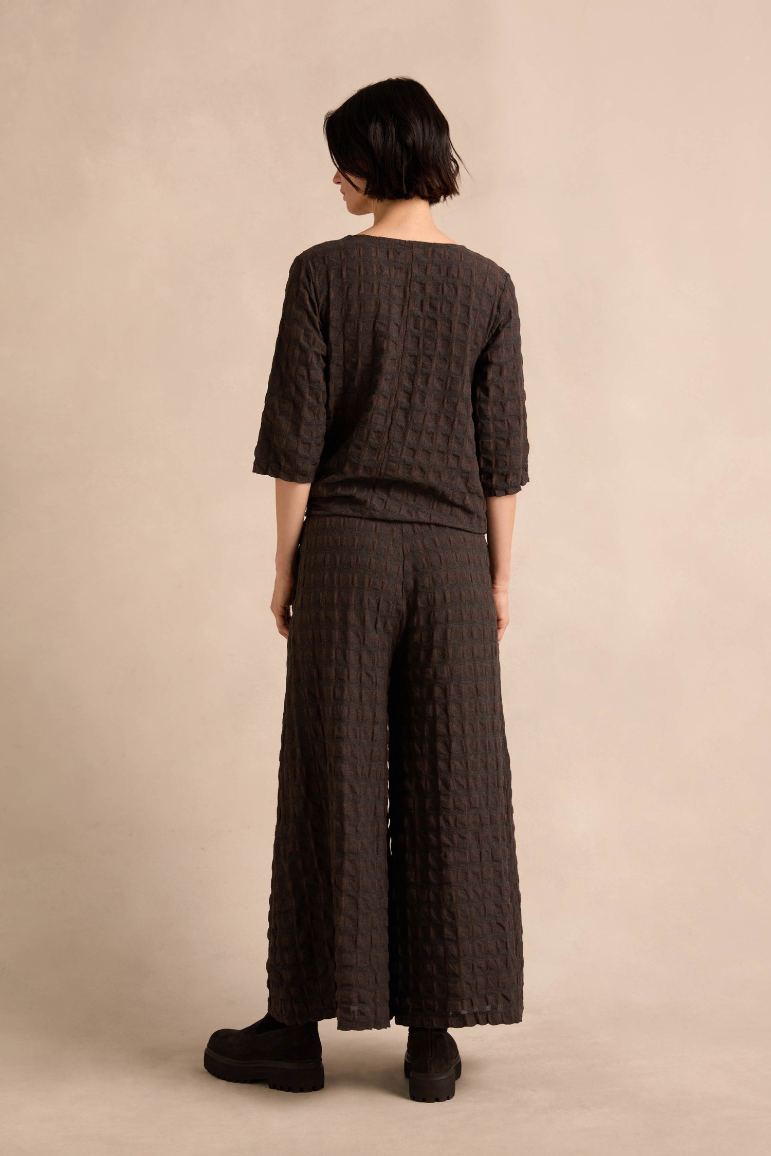 A person stands with their back to the camera, wearing Chocolate Waffle Check Wide Trousers and a matching textured dark top, set against a plain beige background.