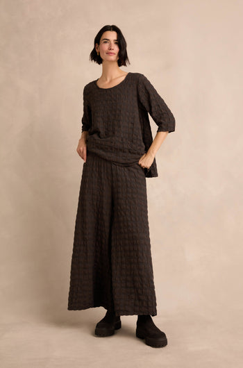 A woman stands against a beige background, wearing the Chocolate Waffle Check Wide Trousers as part of a loose, textured dark two-piece outfit paired with black boots.