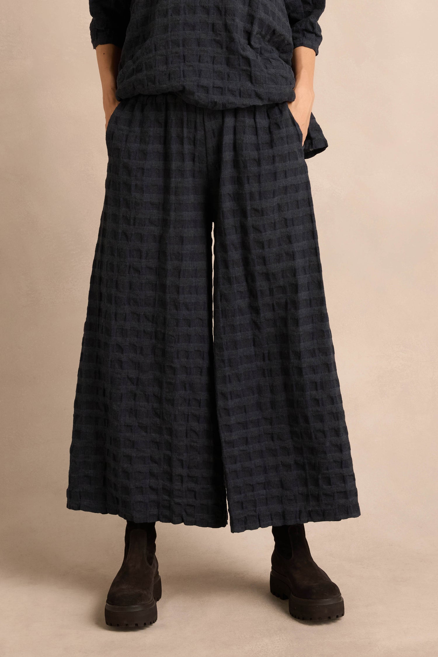 Person wearing loose, wide-leg Navy Waffle Check Wide Trousers crafted from a soft cotton-viscose blend with side pockets, matching top, and black suede ankle boots, standing against a neutral background.