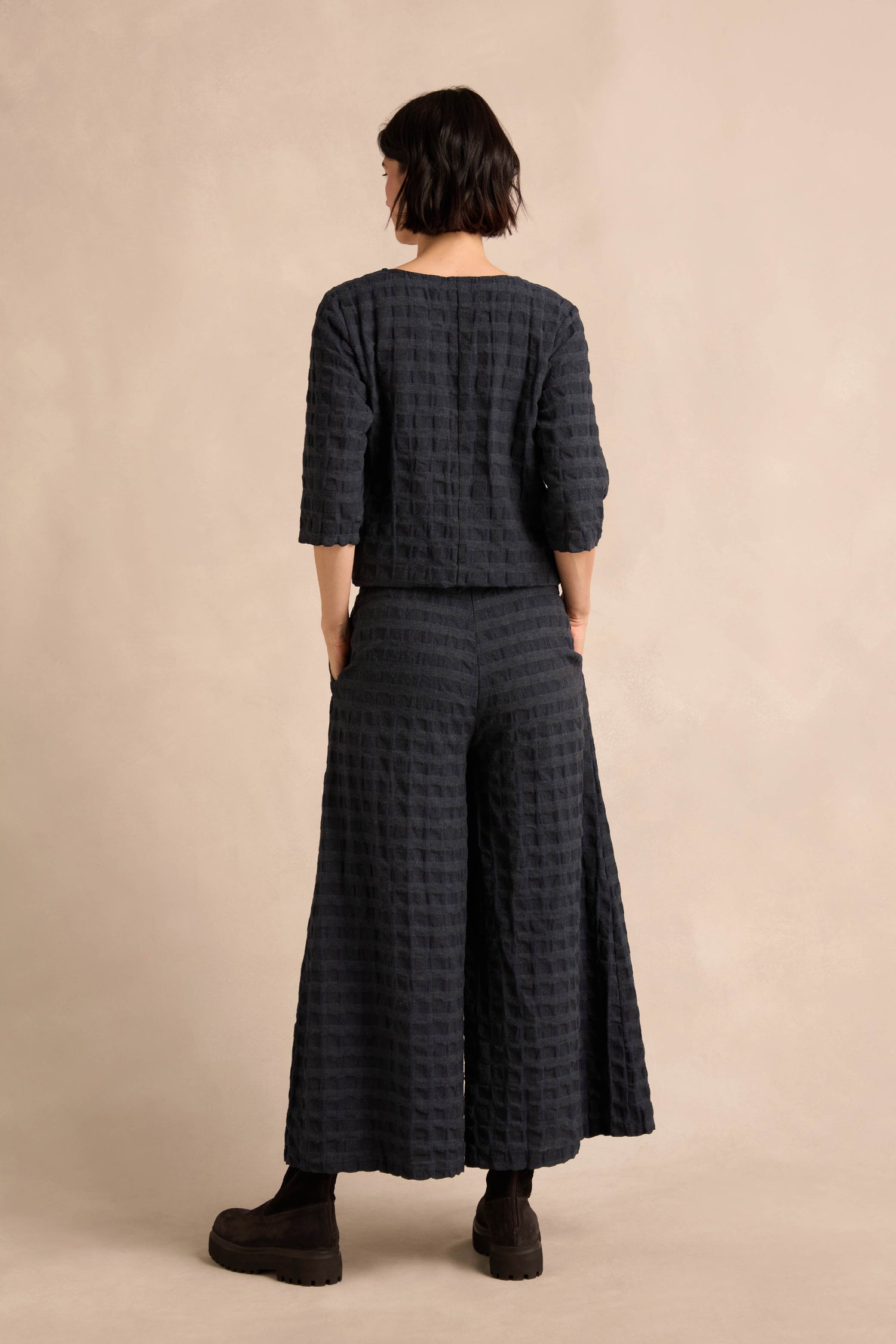 A person stands with their back to the camera, wearing Navy Waffle Check Wide Trousers, paired with a textured dark cotton-viscose top and ankle boots against a neutral background.