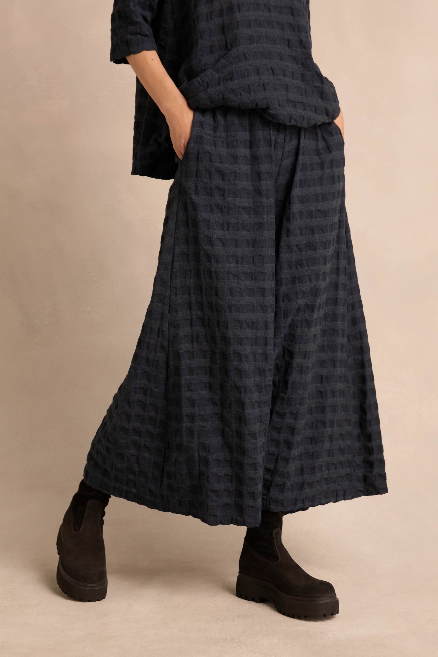 Wearing Navy Waffle Check Wide Trousers, a matching top, and black ankle boots, the person poses with hands in pockets against a neutral background.