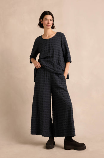 A person stands against a neutral background, wearing a loose-fitting, textured dark top paired with Navy Waffle Check Wide Trousers and chunky black shoes, posing with one hand in their pocket.