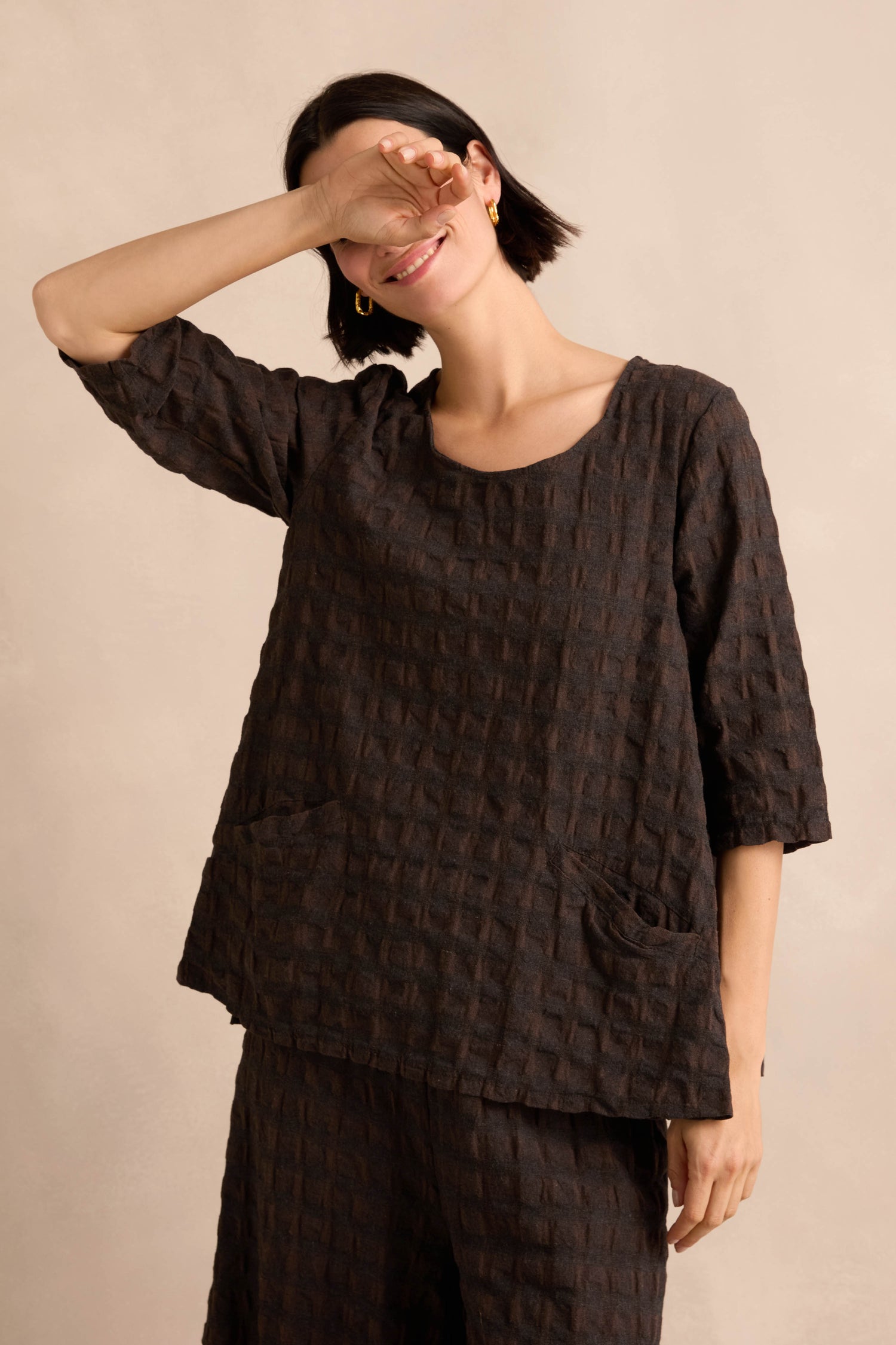 Wearing the Chocolate Waffle Check Flared Top with matching pants, a person stands against a beige background, smiling and raising one arm to partially cover their face.