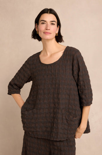 A woman with short dark hair wears the Chocolate Waffle Check Flared Top, featuring three-quarter sleeves, two front pockets, and a loose, textured design crafted from a soft cotton-viscose blend, as she stands against a neutral background.