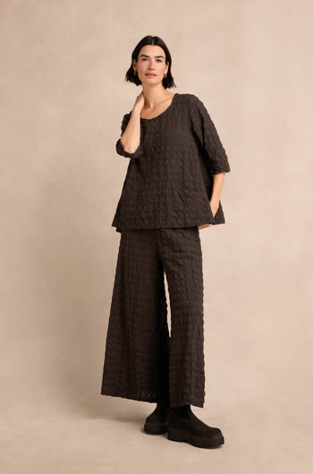 A person stands against a neutral background wearing the Chocolate Waffle Check Flared Top—a loose, textured, dark flared top in cotton-viscose—paired with wide-leg pants and black platform shoes.