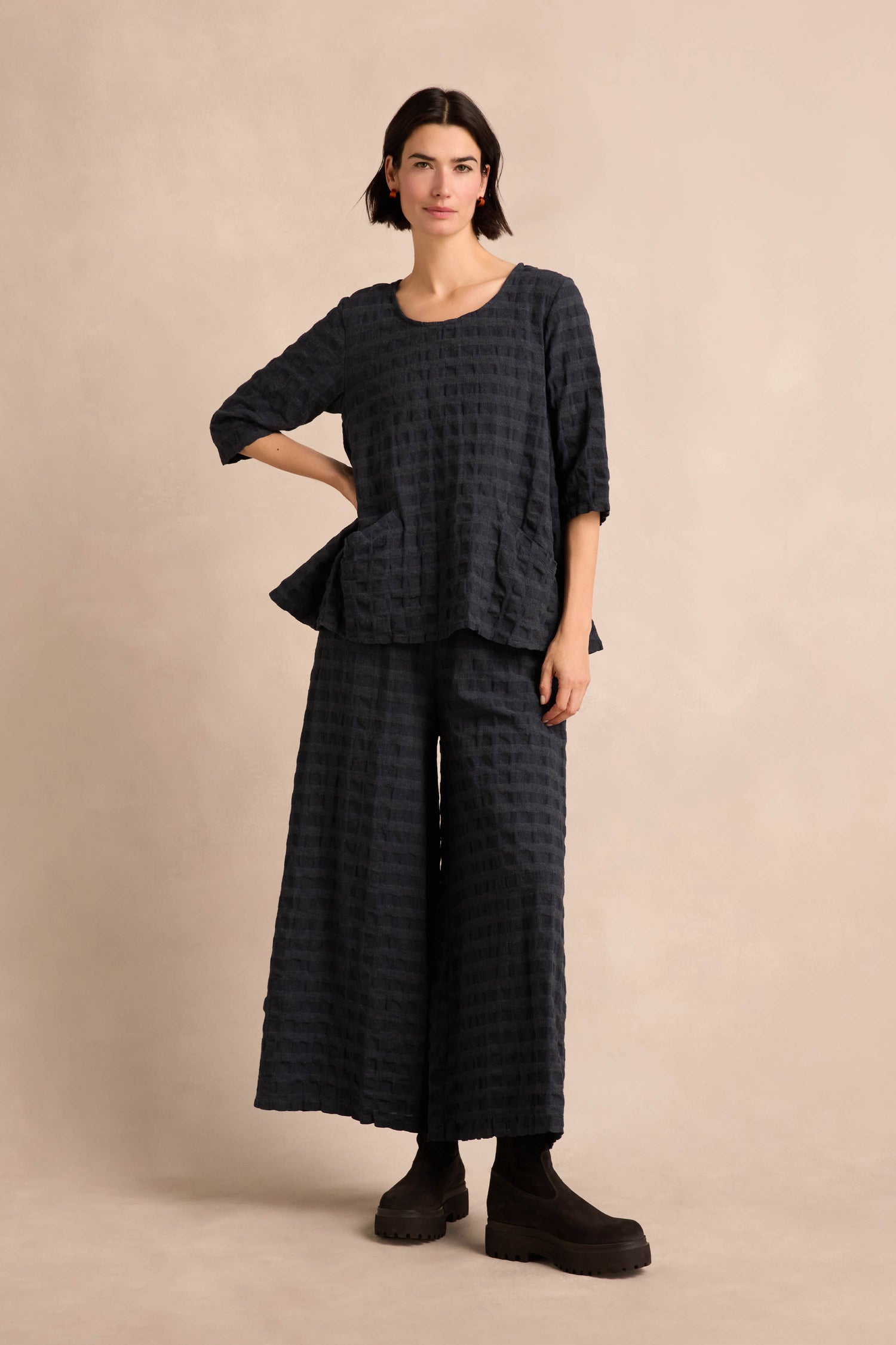 A woman stands against a beige background, wearing the Navy Waffle Check Flared Top—a loose-fitting, textured black top with three-quarter sleeves, crafted from a soft cotton-viscose blend—paired with matching wide-leg pants and black shoes.