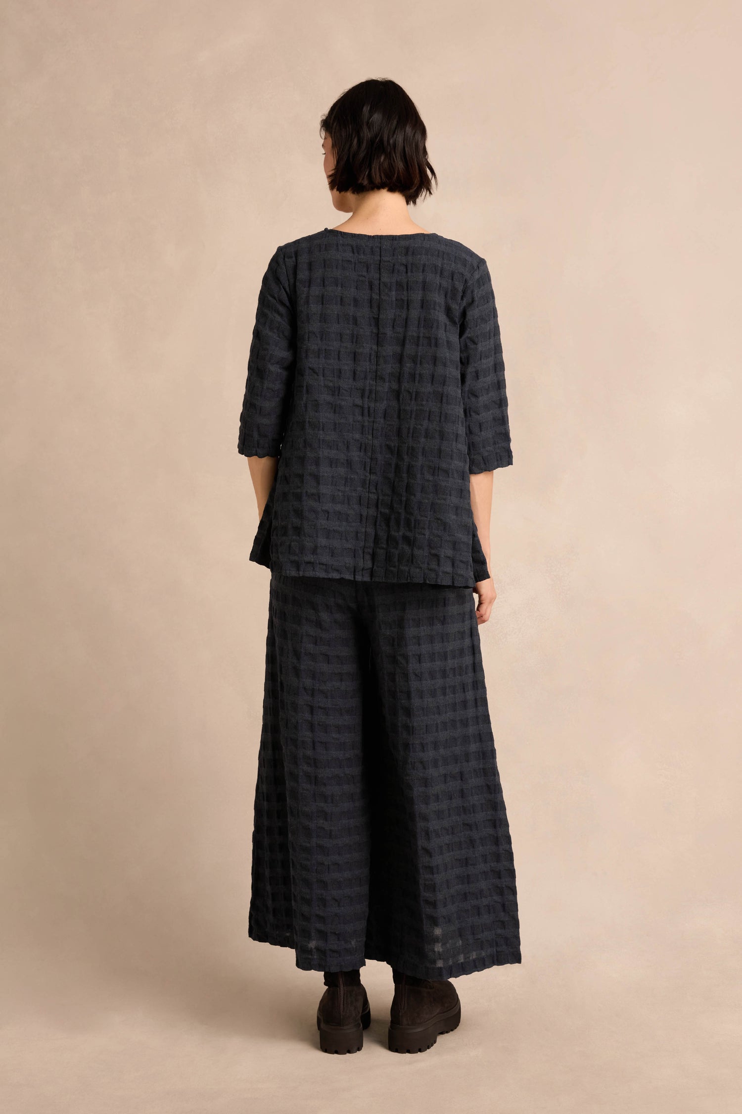 A person stands facing away, wearing the Navy Waffle Check Flared Top—a loose-fitting, textured black top with elbow-length sleeves—paired with matching wide-leg pants, both crafted from soft cotton-viscose, against a neutral background.