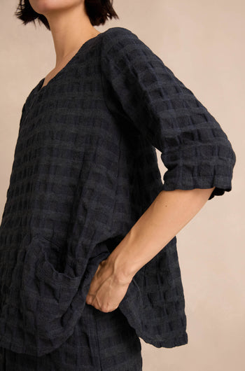 A person wearing the Navy Waffle Check Flared Top, featuring a loose, textured design with three-quarter sleeves, stands with one hand in the pocket against a neutral background.