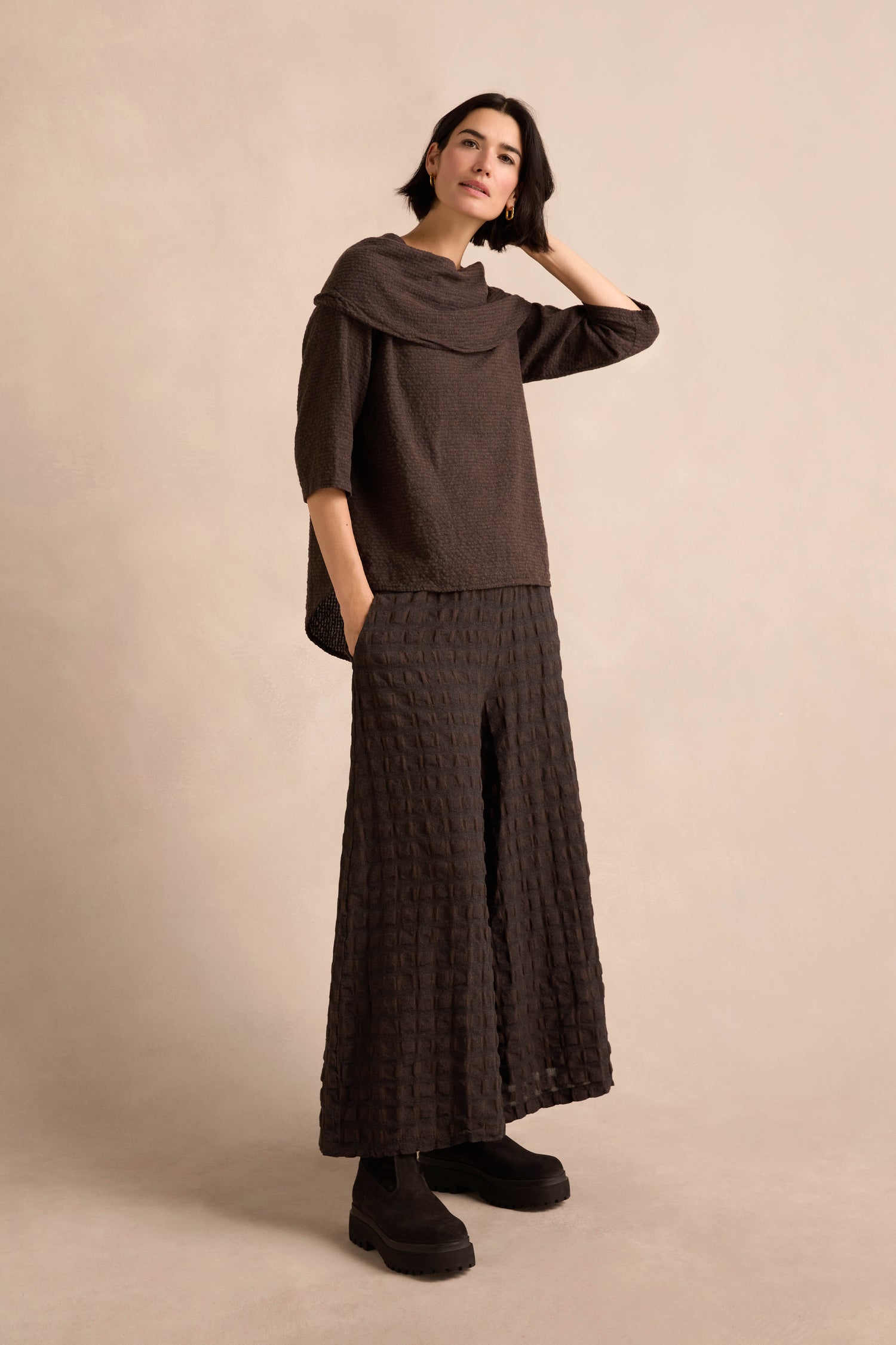 A woman stands against a beige backdrop, wearing the Chocolate Draped Neck Top paired with matching wide-leg pants and black platform shoes, with one hand touching her head.