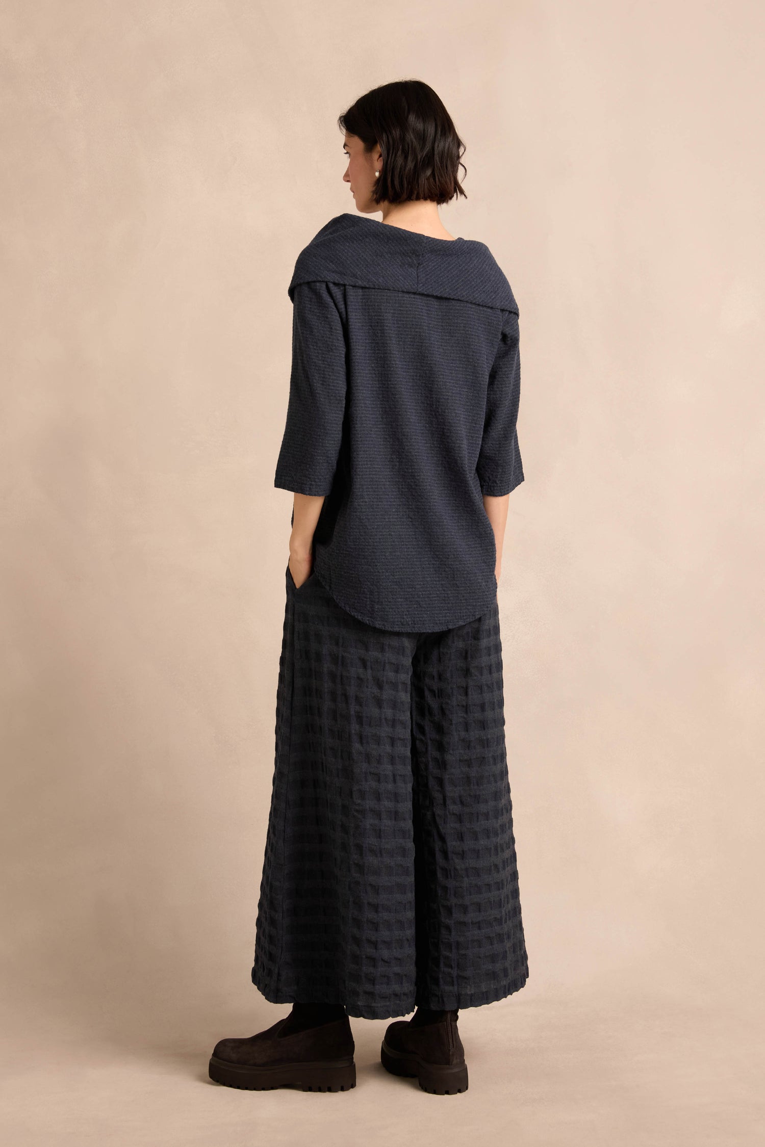 A person stands facing away, wearing the Navy Draped Neck Top—a relaxed-fit, navy blue piece with a wide collar—paired with textured navy wide-leg pants and black platform shoes, against a beige background.