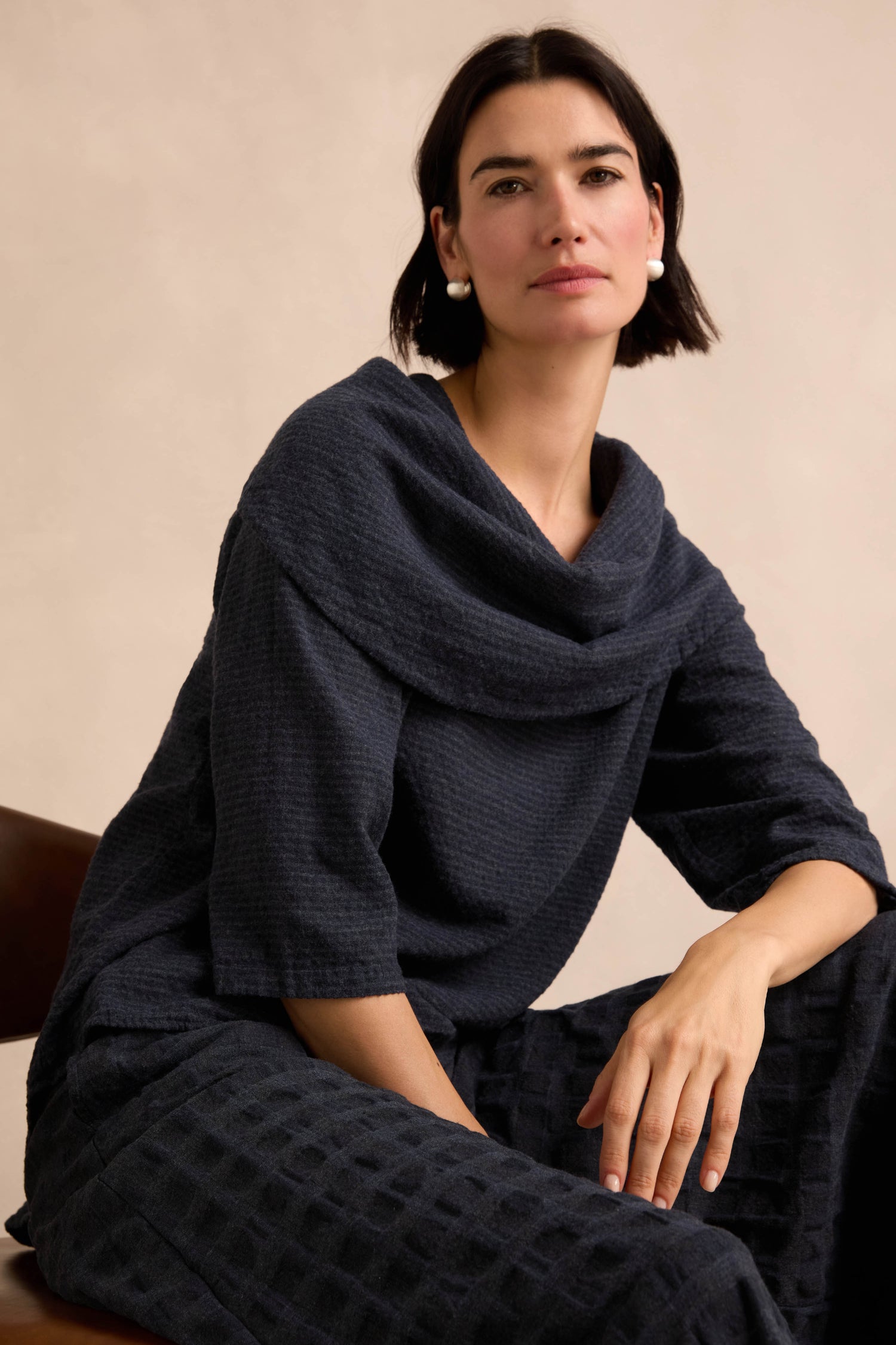 A woman with short dark hair sits in a chair wearing the Navy Draped Neck Top and pearl earrings, looking ahead with a neutral expression against a beige background.