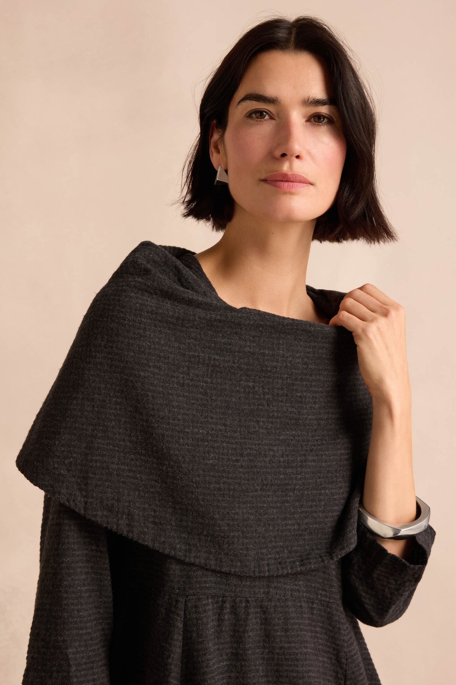 A woman with short dark hair is looking at the camera, holding the edge of her Anthracite Draped Neck Bubble Dress, which features a textured fabric and a draped cowl neck, against a neutral background.