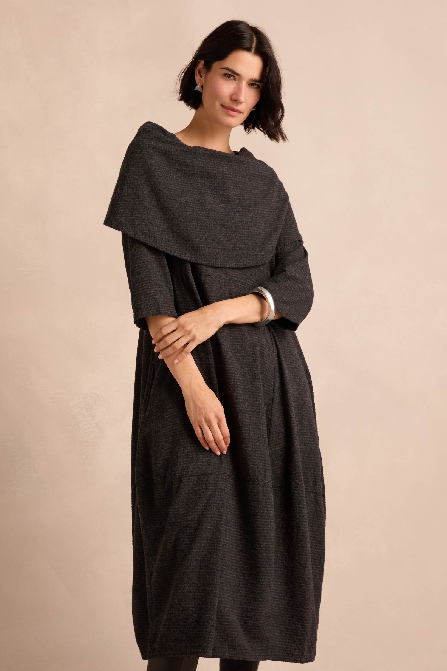A woman stands against a neutral background, wearing the Anthracite Draped Neck Bubble Dress—a loose-fitting design in anthracite grey featuring a wide draped collar and minimal accessories.