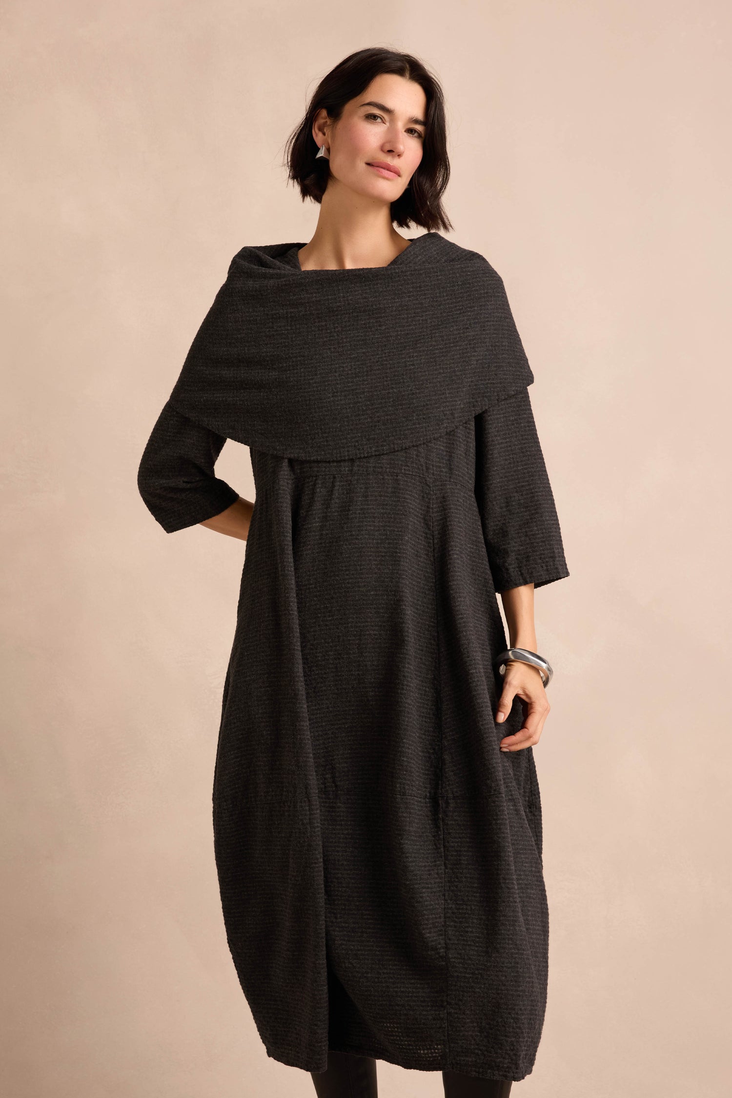 A woman stands against a neutral background wearing the Anthracite Draped Neck Bubble Dress, featuring a textured finish, loose fit, and a wide draped cowl neck.