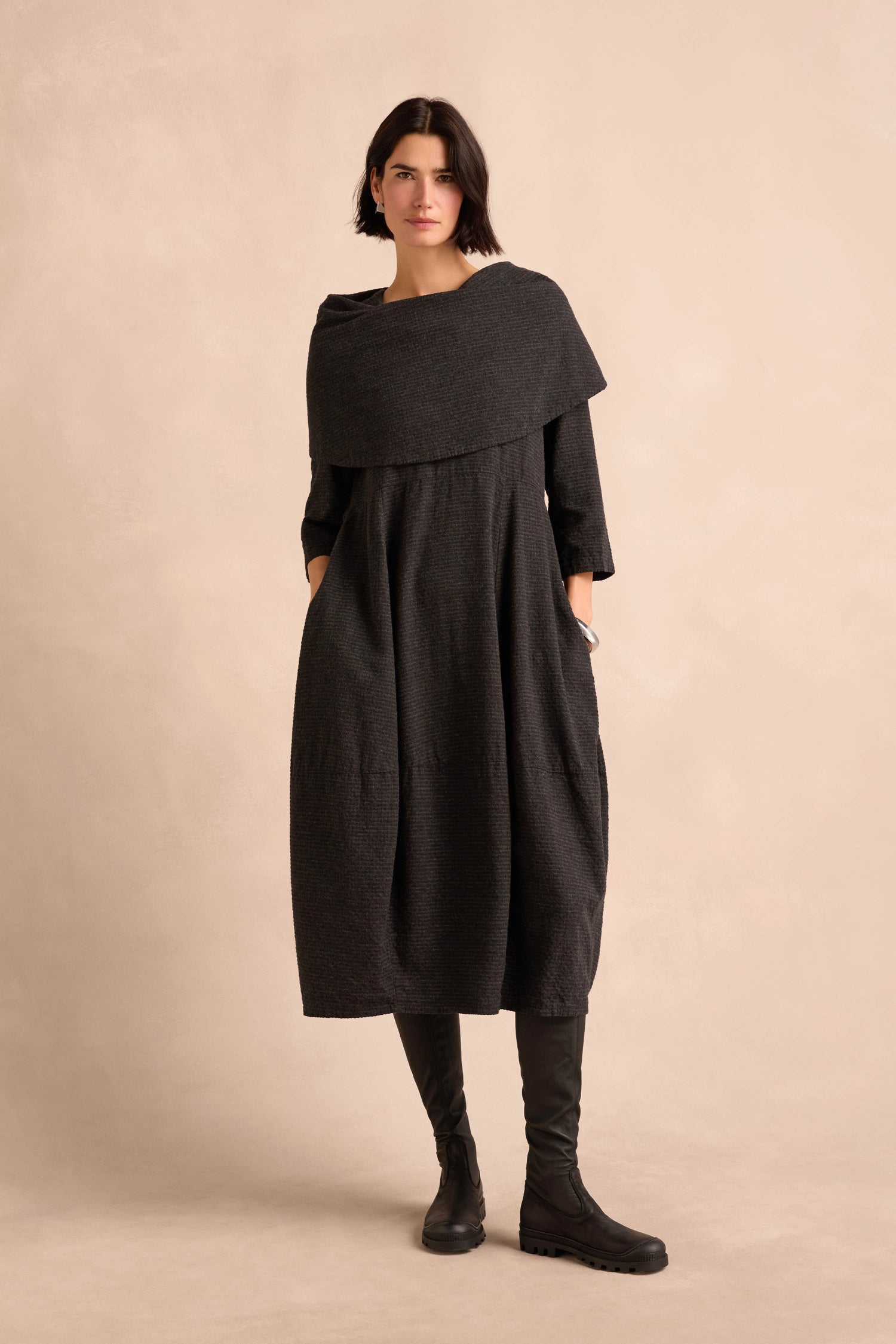 A woman stands against a beige background wearing the Anthracite Draped Neck Bubble Dress, featuring a textured fabric, wide collar, and long sleeves, paired with black boots.
