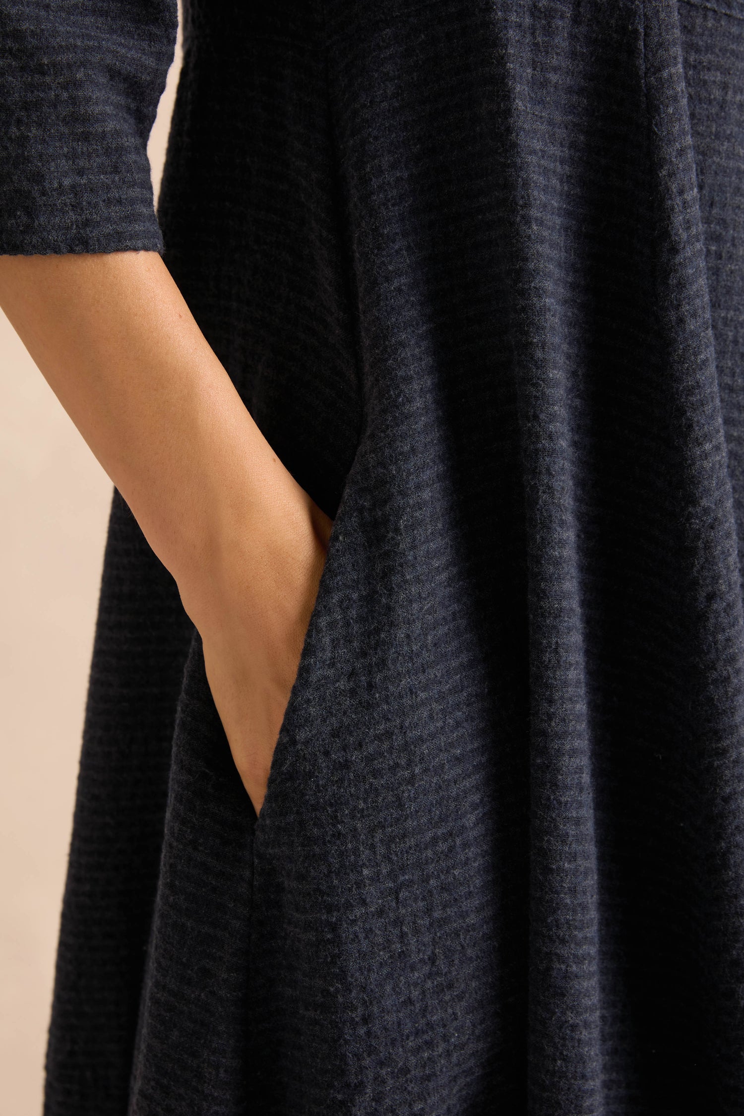 A person’s hand is placed in the pocket of the Navy Draped Neck Bubble Dress, a dark, textured cotton-viscose jersey style with three-quarter sleeves.