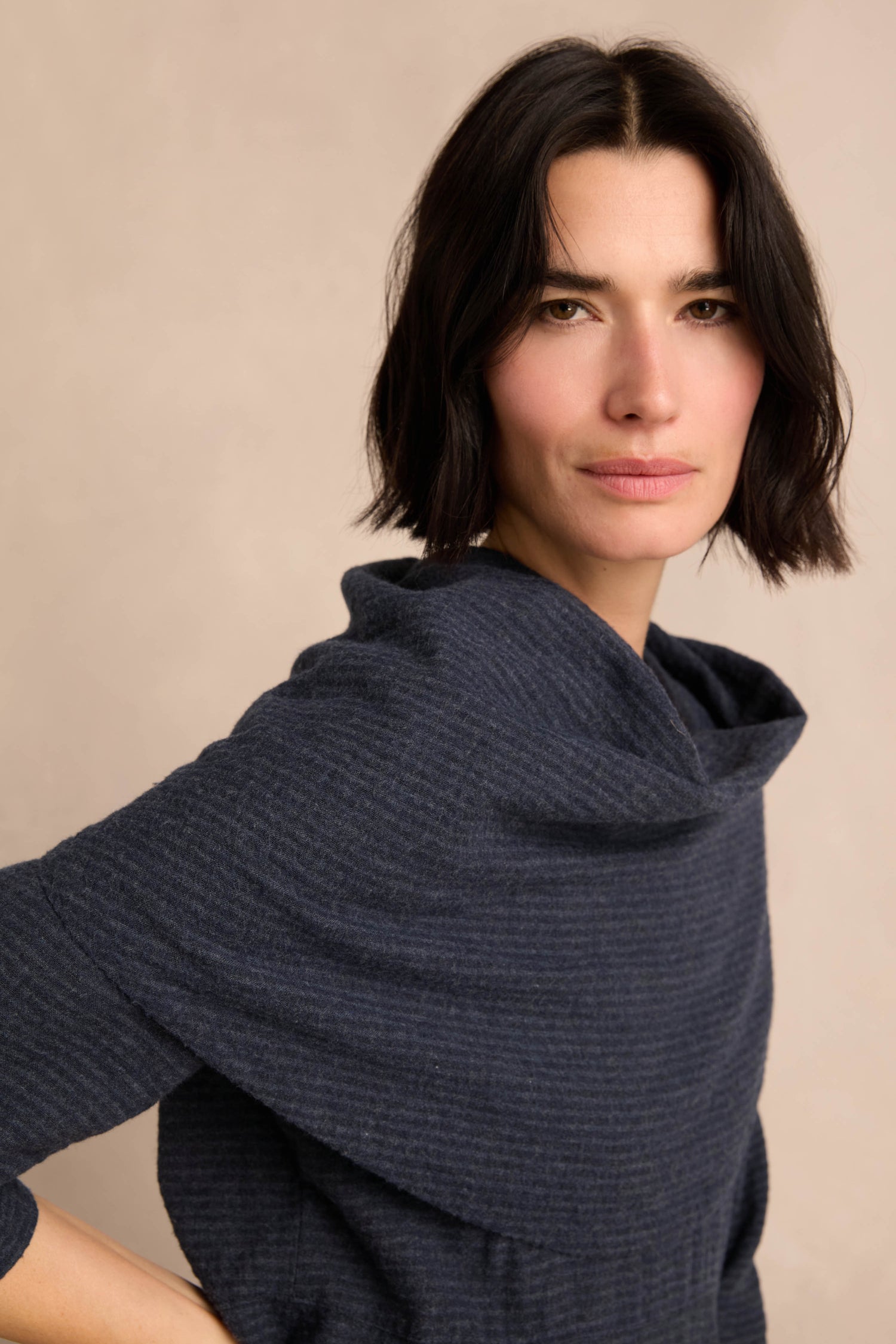 A person with short dark hair and a neutral expression wears the Navy Draped Neck Bubble Dress, standing against a plain beige background.