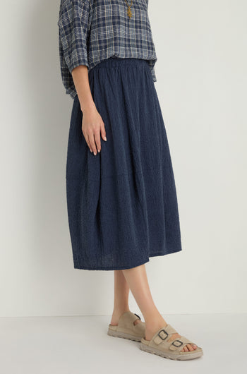 Someone wears a blue plaid shirt, beige sandals, and the Crinkle Bubble Skirt—a navy blue textured cotton-blend skirt with an elastic waist—while standing against a plain light background.