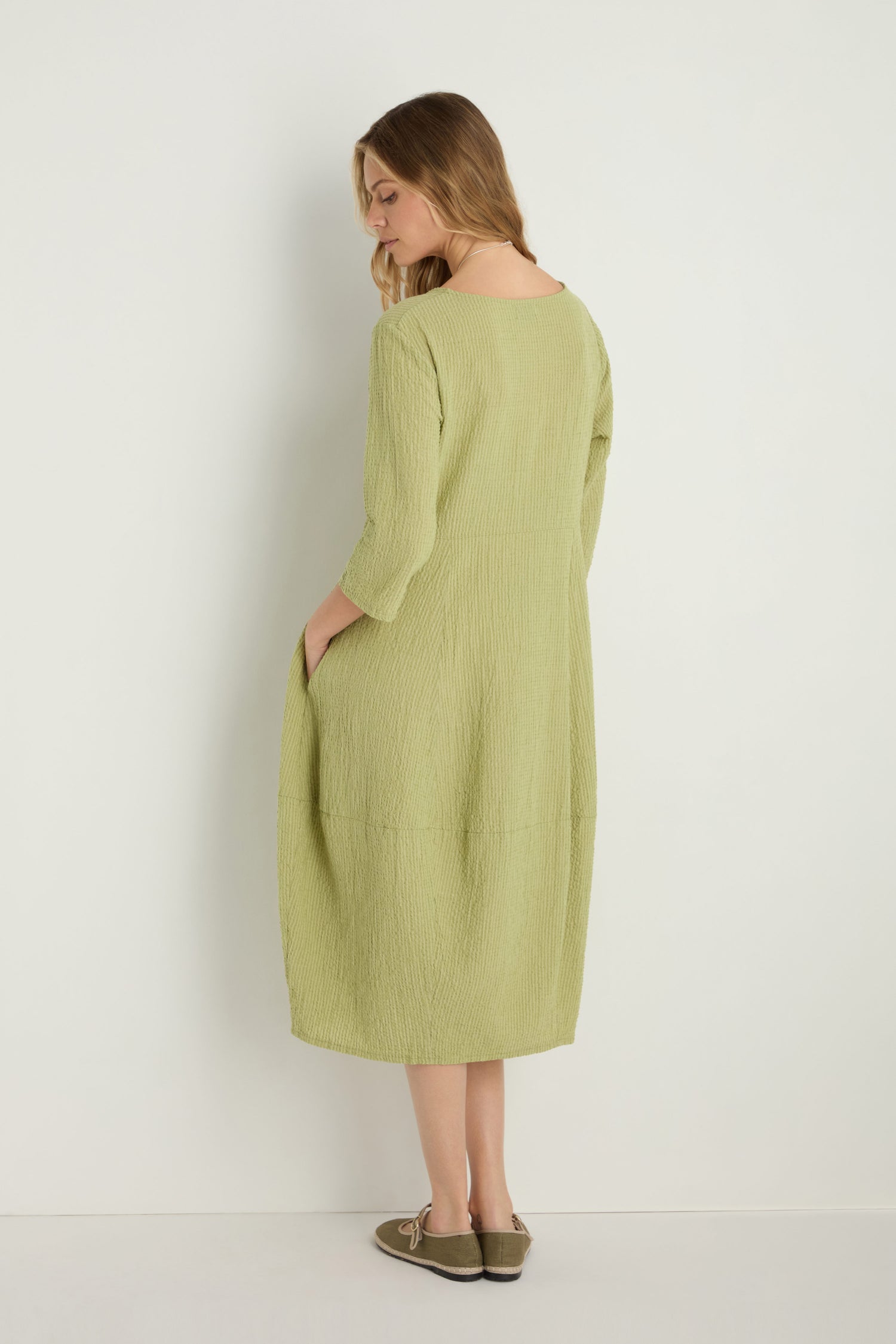 A woman wears the Crinkle Bubble Dress in light green, relaxed fit, hands in pockets, facing away against a plain white background.