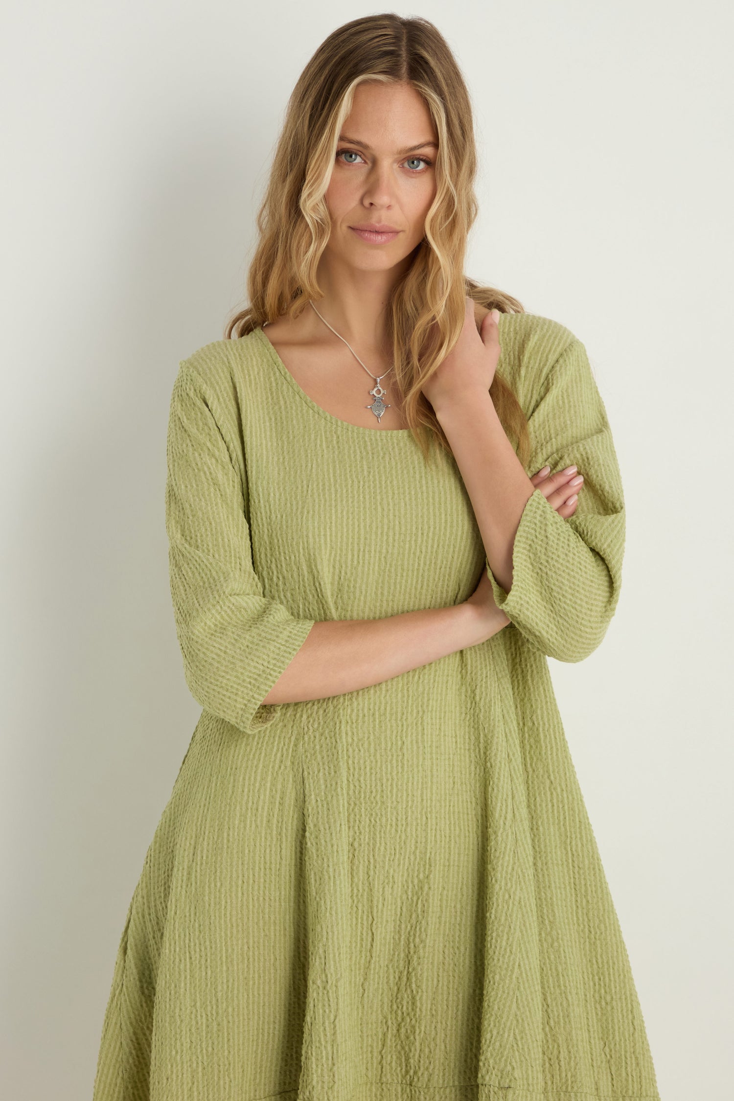 A woman with long wavy blonde hair wears the Crinkle Bubble Dress in light green, made from a textured cotton blend, standing against a plain background and looking at the camera with her arms gently crossed.