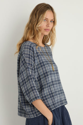 A woman with wavy blonde hair models the Check Boxy Top with a long necklace and a navy textured skirt, standing against a plain light background.