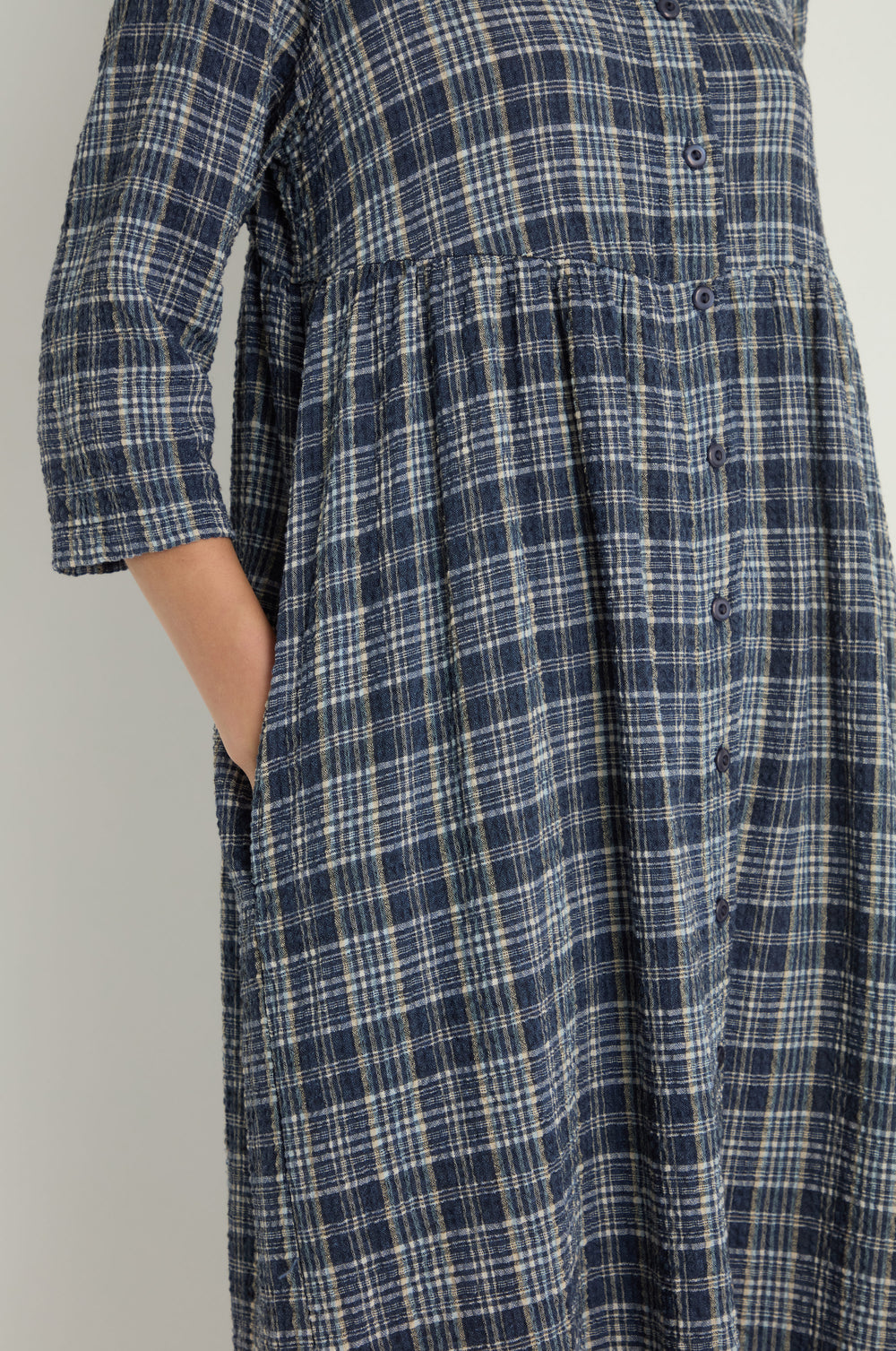 A person wears the Check Button-Through Navy Dress, featuring a blue and white plaid pattern, three-quarter sleeves, a pocket detail, and classic Danish design.
