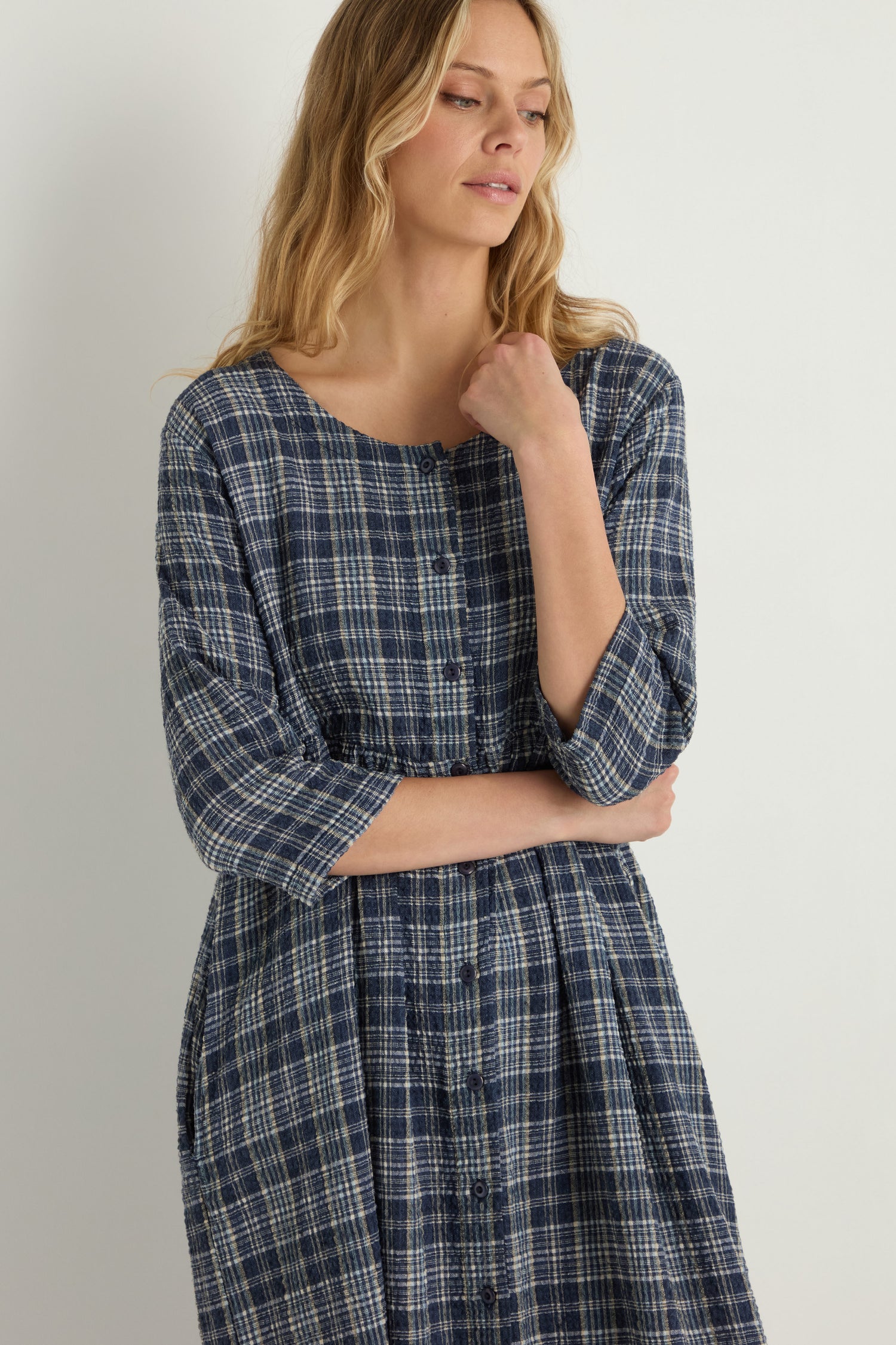 A woman stands against a plain white background wearing the Check Button-Through Navy Dress, looking to the side with one hand near her chin, reflecting timeless Danish design.