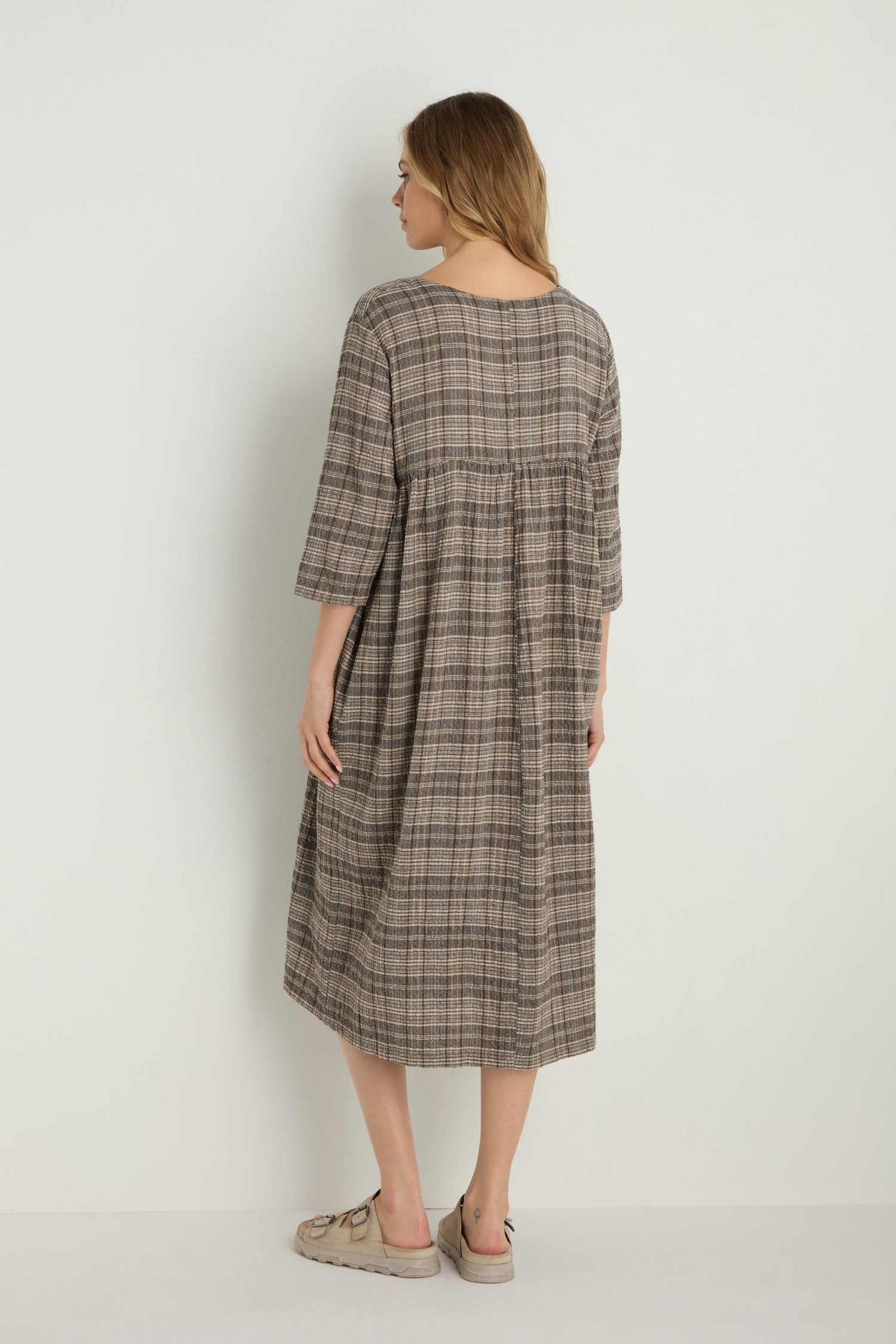 A woman faces away, wearing the Check Button-Through Taupe Dress—a loose-fitting, knee-length plaid dress with three-quarter sleeves—paired with light sandals against a plain background.