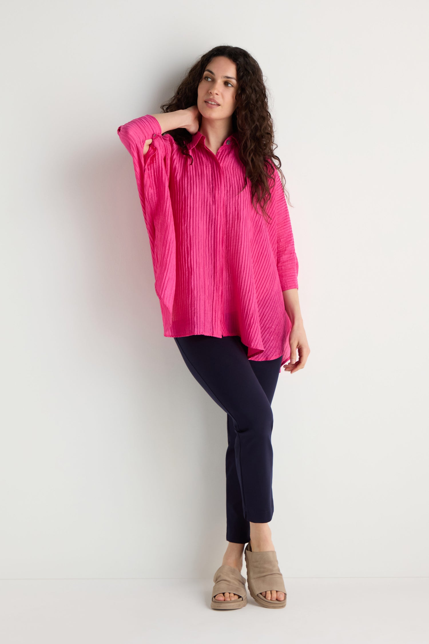 A person stands against a white wall wearing the Pleated Easy Shirt in bright pink, navy pants, and beige sandals.