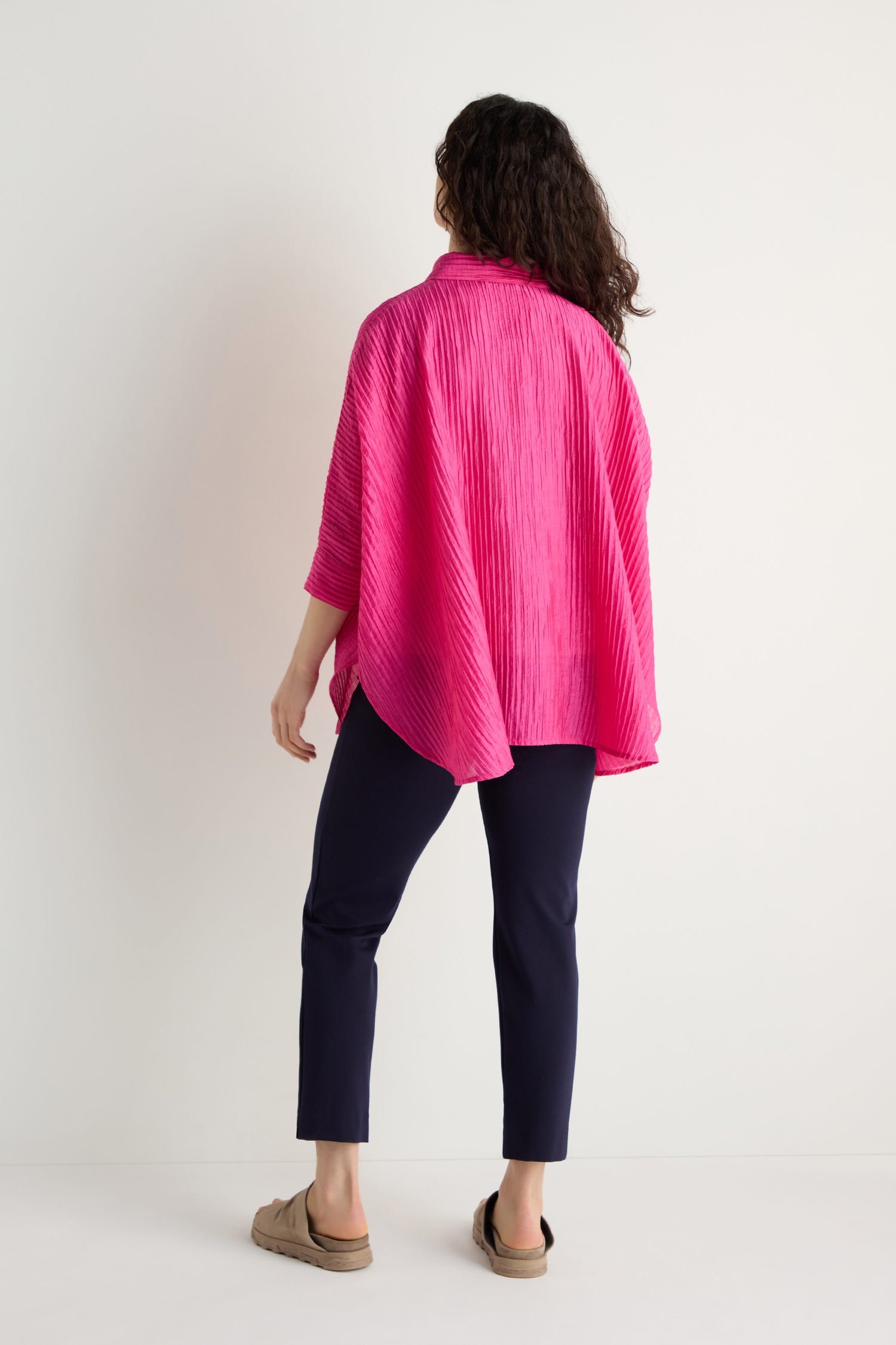 A person faces away in a bright pink Pleated Easy Shirt, paired with dark pants and beige slip-on shoes, set against a plain light background.