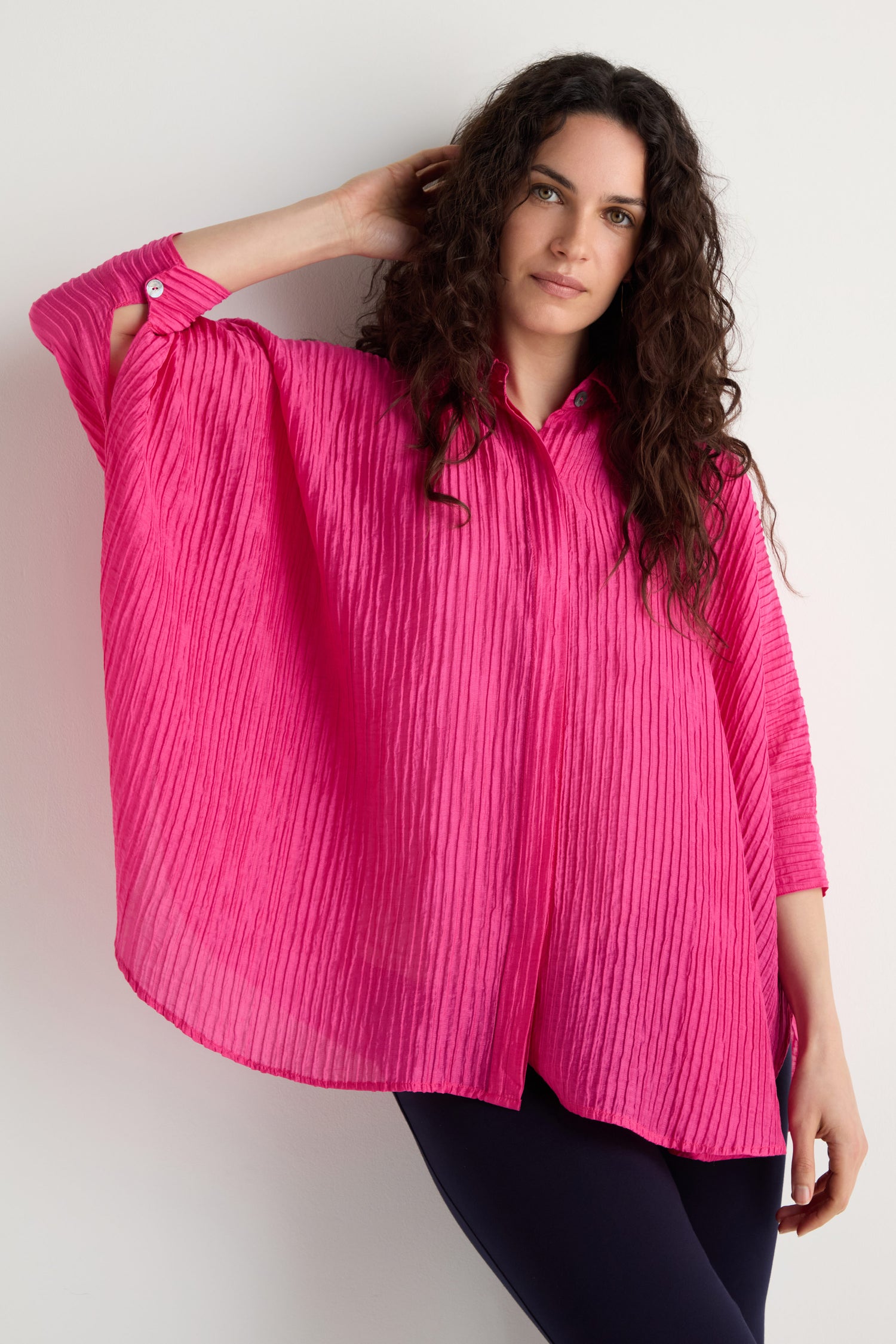 A woman with long curly hair wears the Pleated Easy Shirt in bright pink and dark pants, standing against a white background.