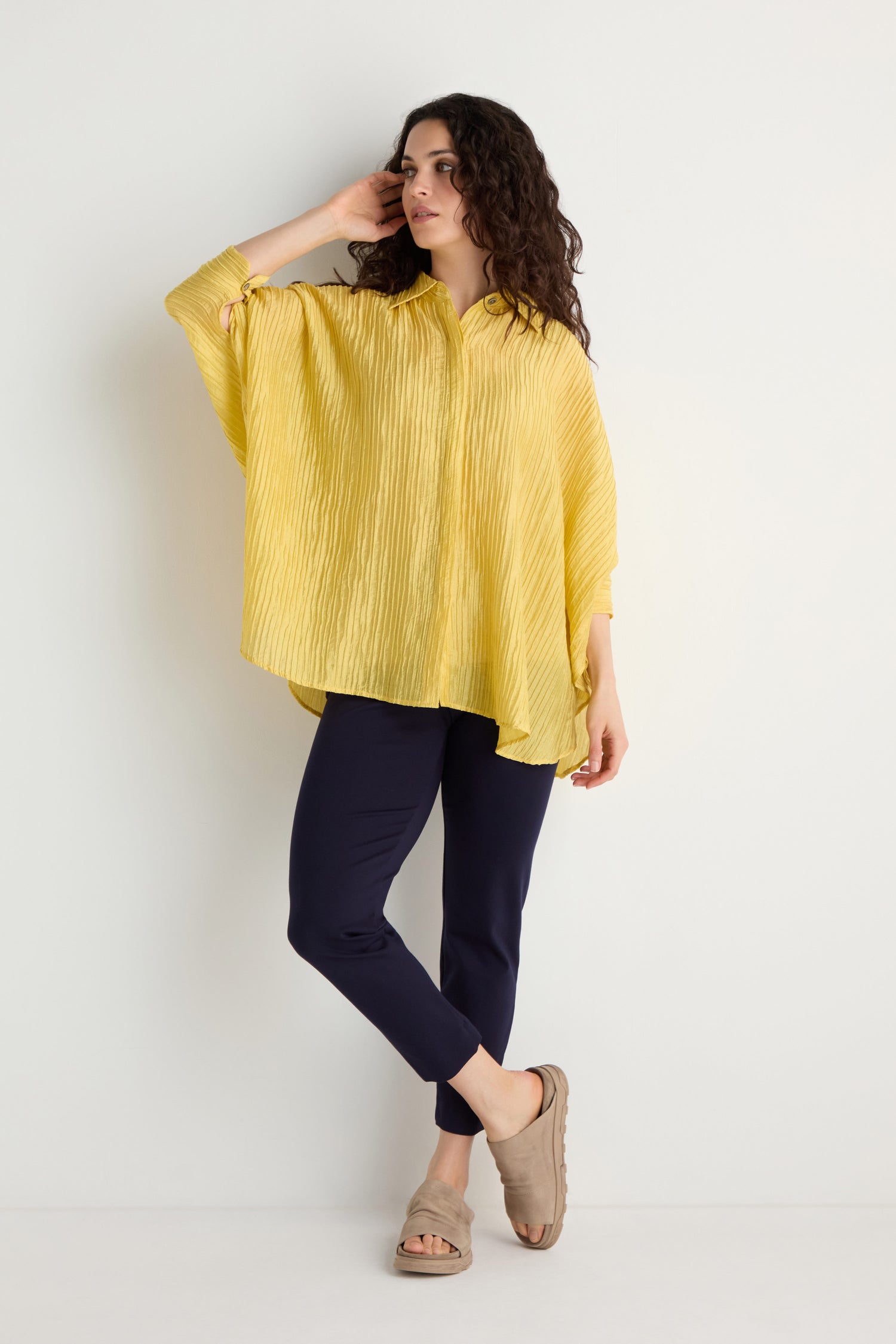 A woman models the Pleated Easy Shirt in yellow, paired with navy pants and beige sandals, standing against a plain white background.