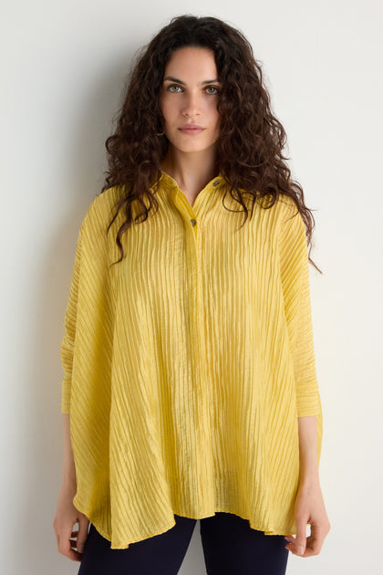 Woman with long curly hair wears the Pleated Easy Shirt, an oversized textured yellow blouse, paired with dark pants against a plain white background.