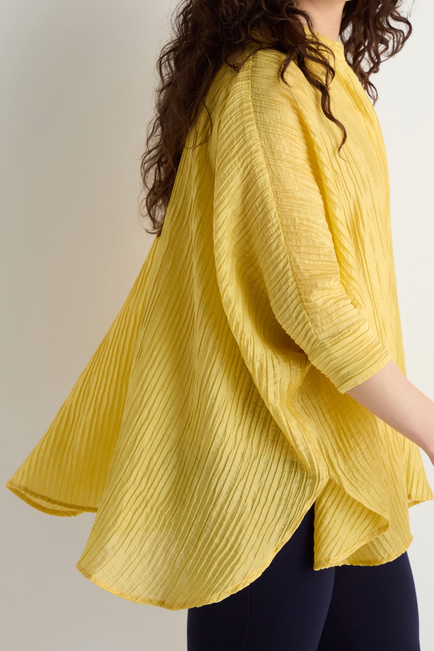 A person shown from the side wearing the Pleated Easy Shirt, a loose-fitting, pleated yellow oversized top with three-quarter sleeves, paired with black pants.