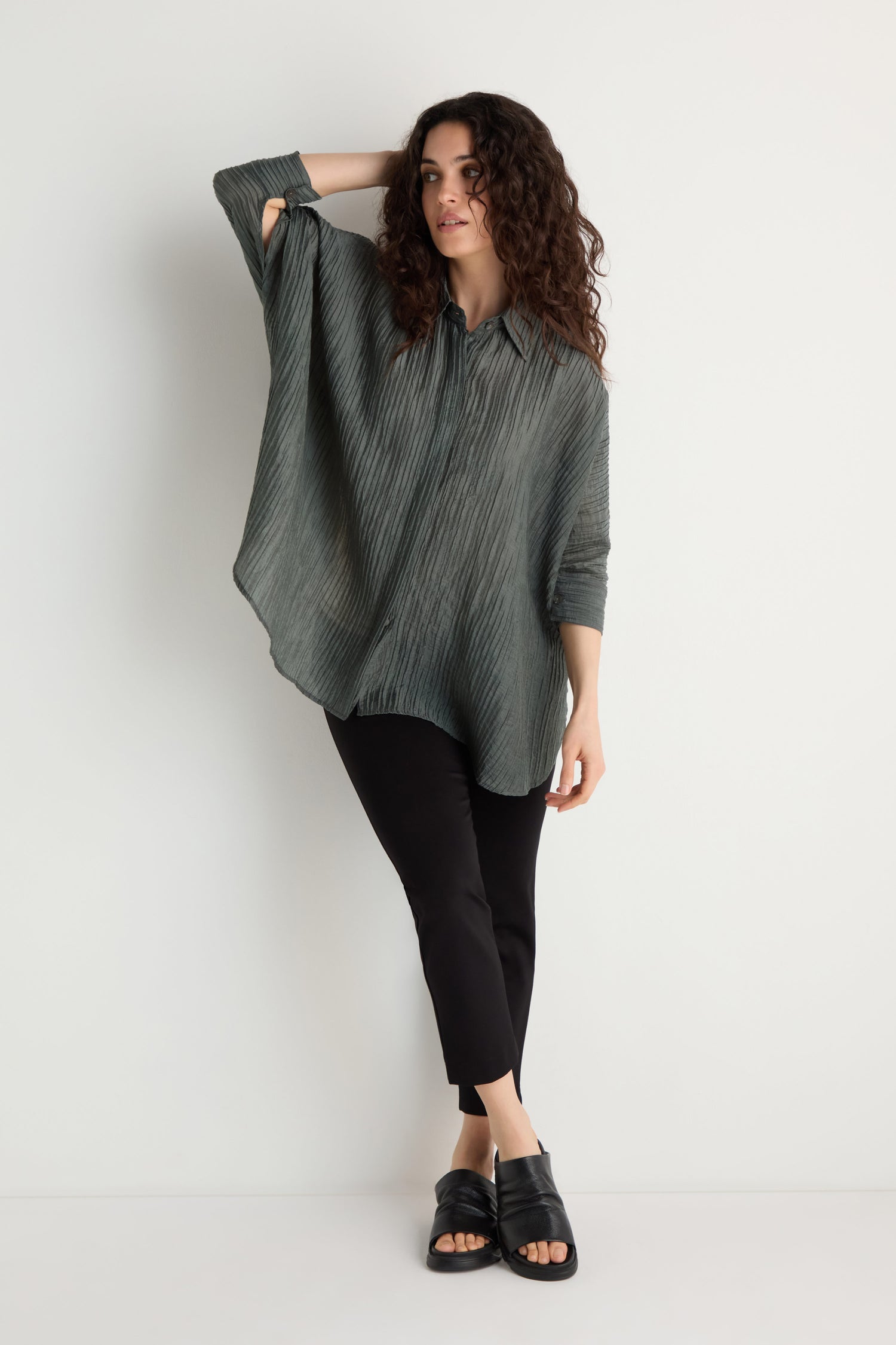 A woman stands against a white wall wearing the Pleated Easy Shirt in gray, paired with black pants and black sandals.
