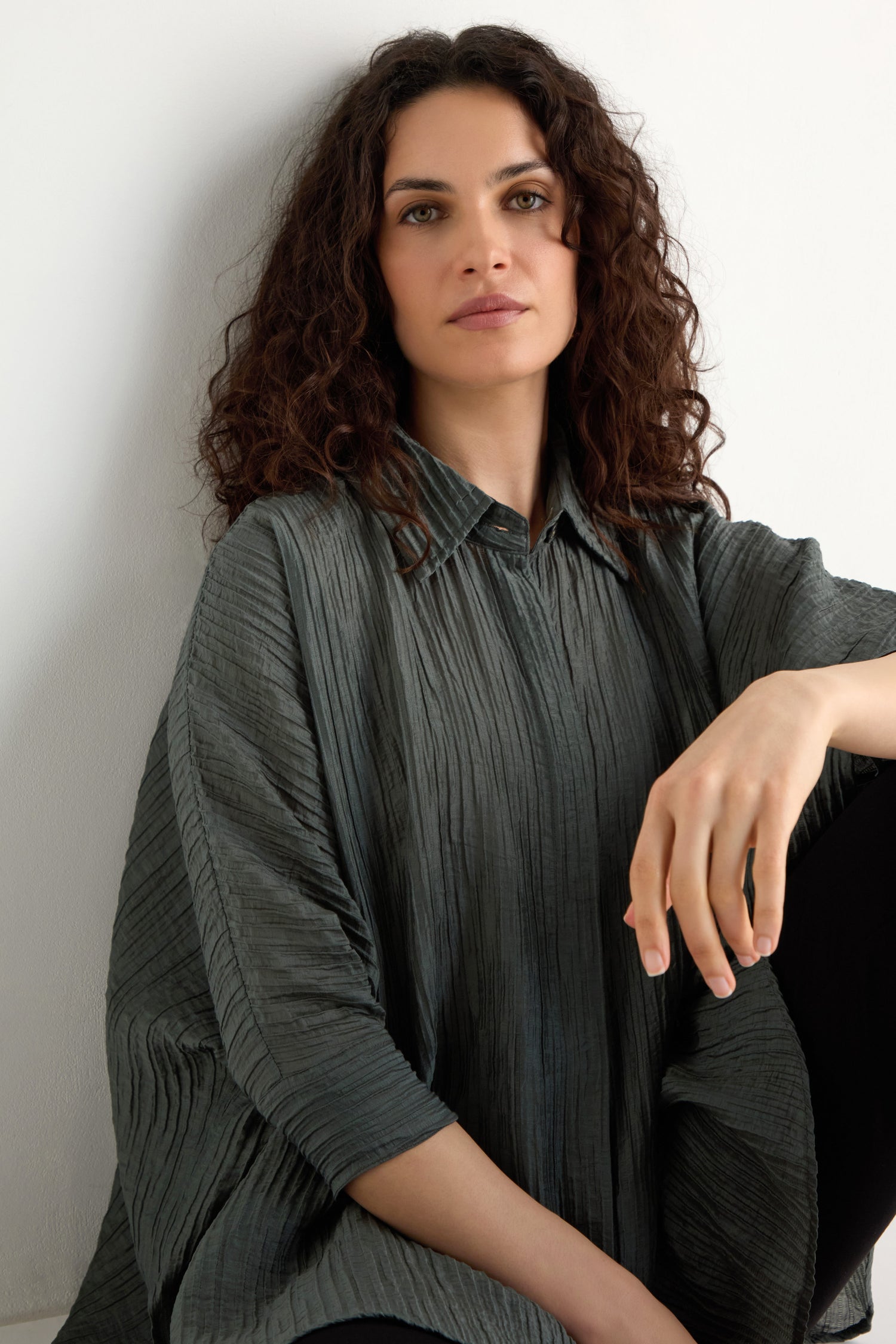 A woman with curly brown hair sits against a white wall, wearing the Pleated Easy Shirt in gray, looking relaxed.