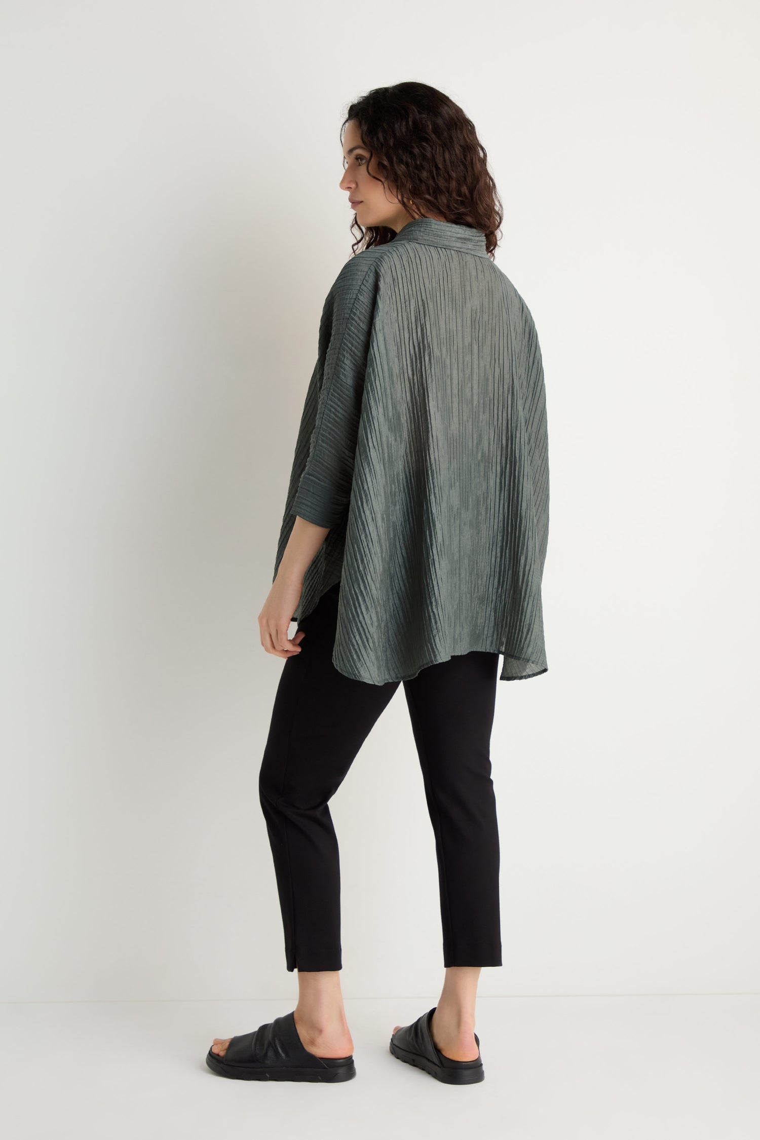 A woman stands sideways against a white background, wearing the dark green Pleated Easy Shirt with black pants and black slide sandals.